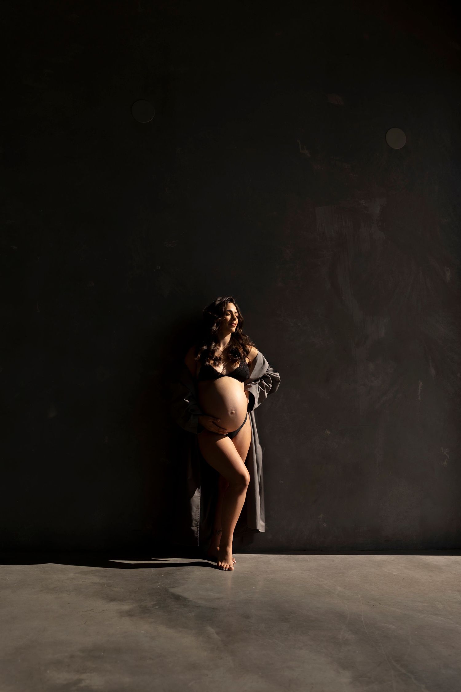 Pregnant woman in beige lingerie and sheer robe cradling baby bump against dark studio backdrop.