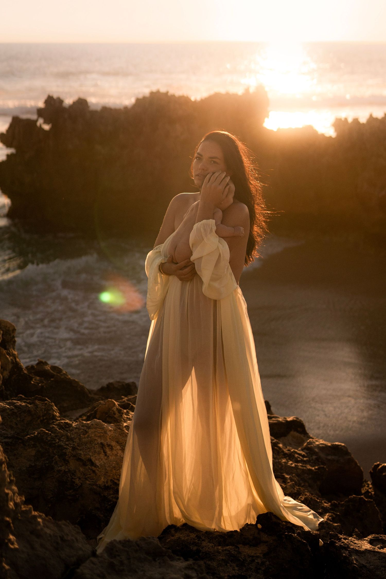 Woman in flowing cream gown stands on rocky beach at golden sunset, ocean waves glimmering behind her.