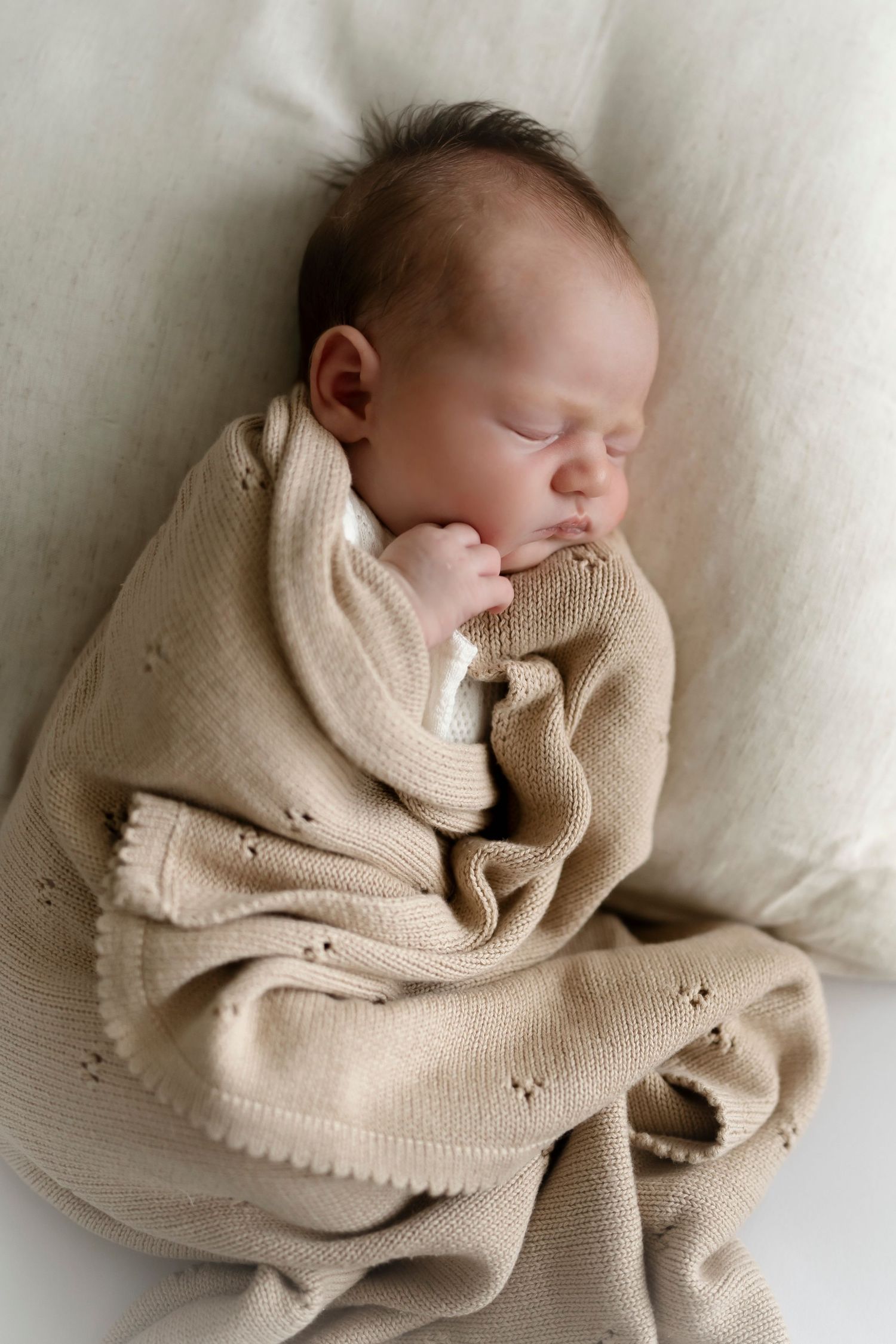 Peaceful newborn baby sleeping soundly, swaddled in a soft beige knit blanket on a white cushion.