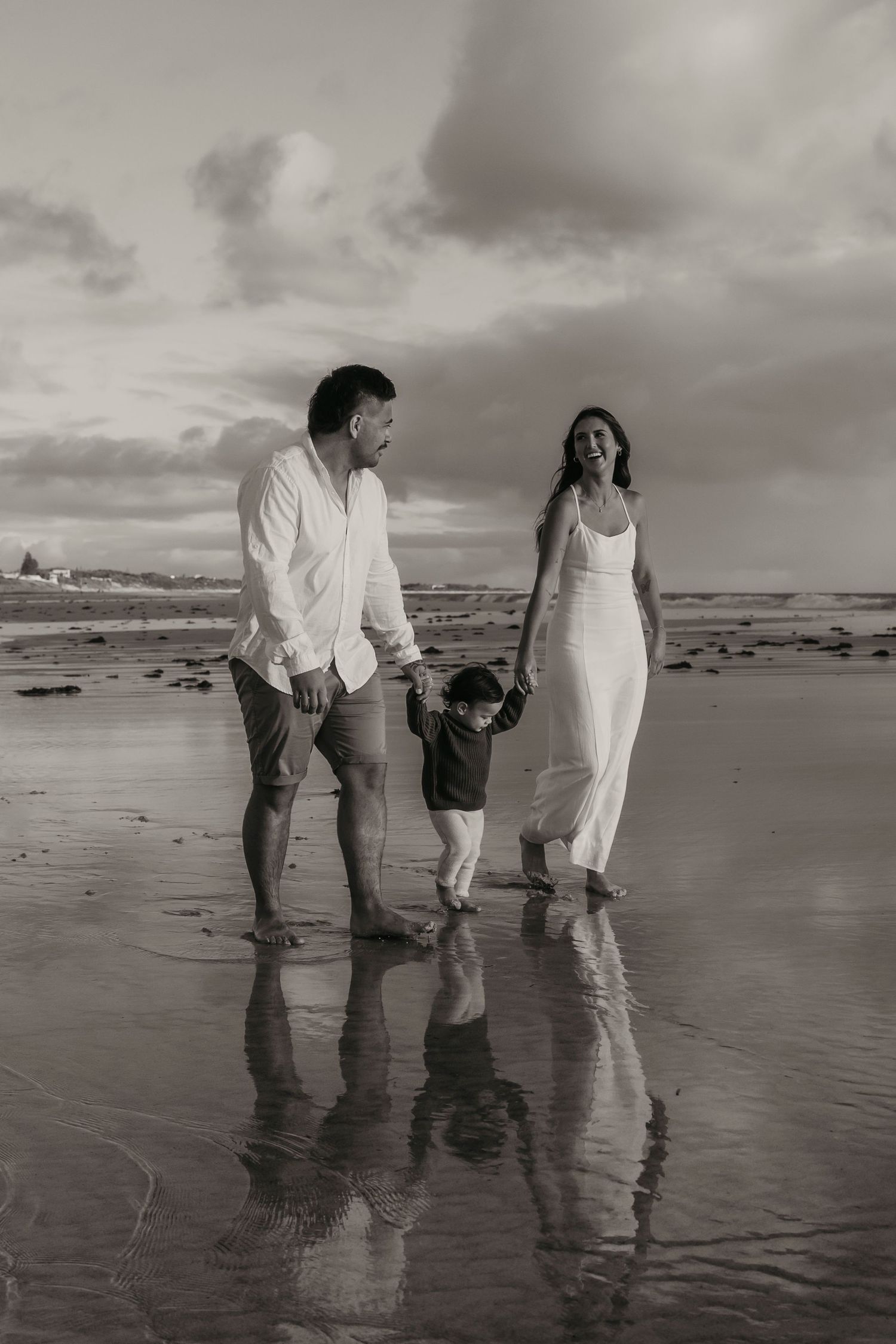 Family walking together along a reflective beach shoreline at sunset in black and white.