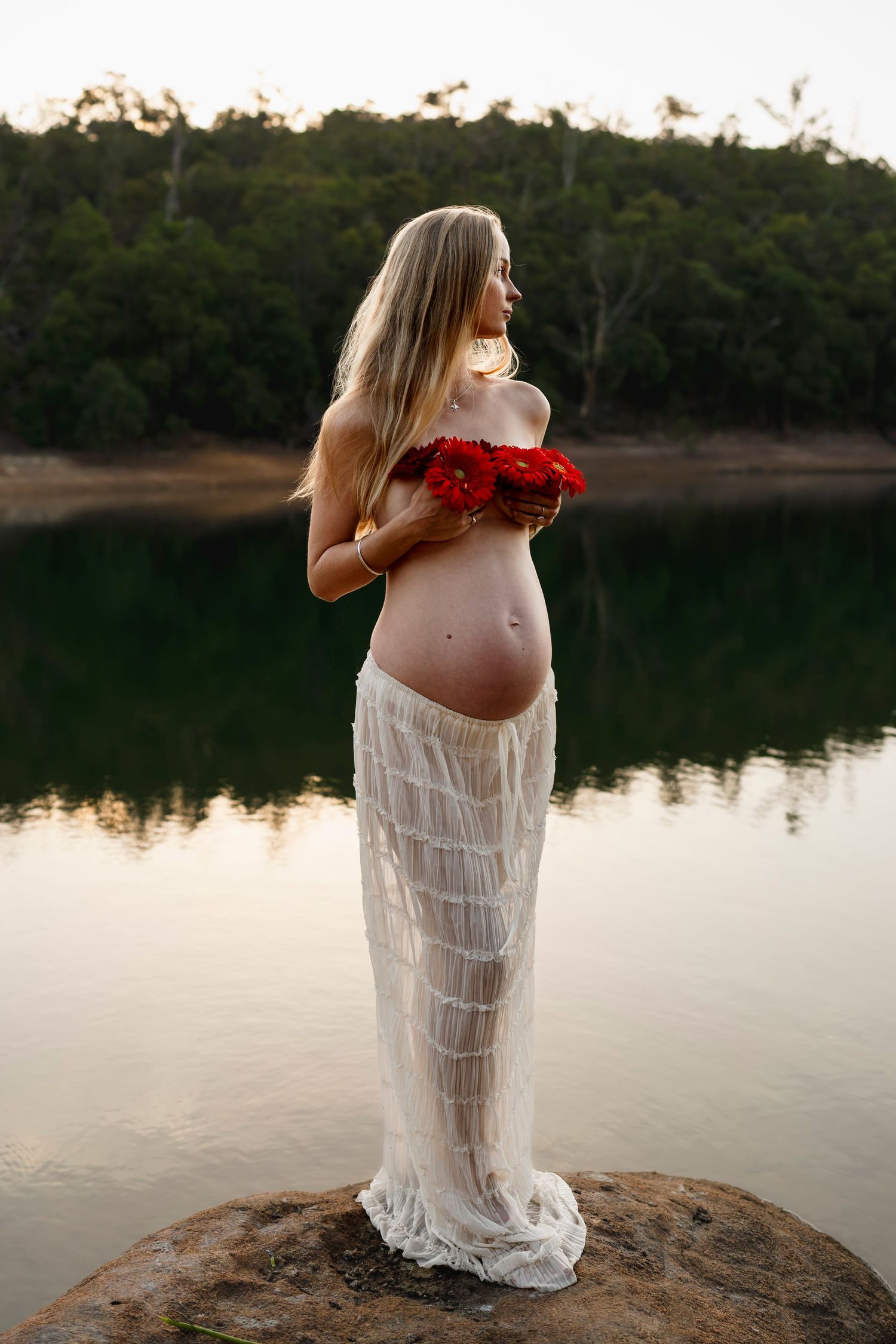 A maternity photo at sunset features an expectant mother in a sheer white dress holding red roses by a tranquil lake.