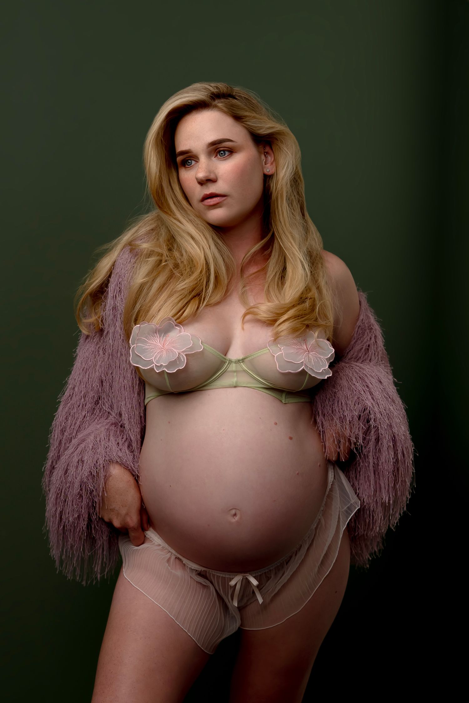 Pregnant woman in lingerie and pink fuzzy jacket posing against dark green background.