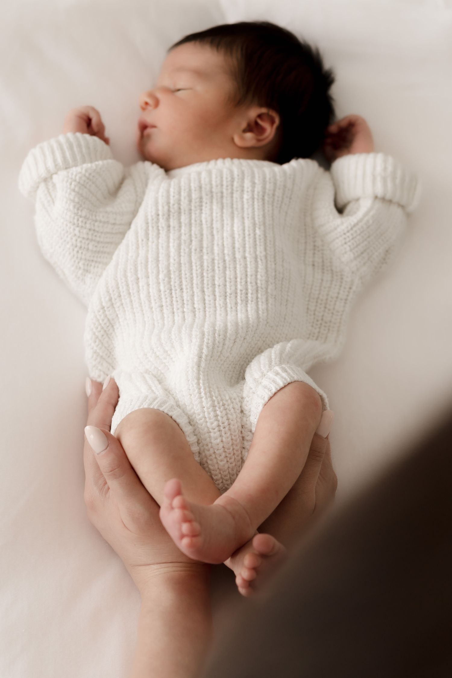 A sleeping newborn baby wearing a cozy white knit romper rests peacefully on soft white fabric.