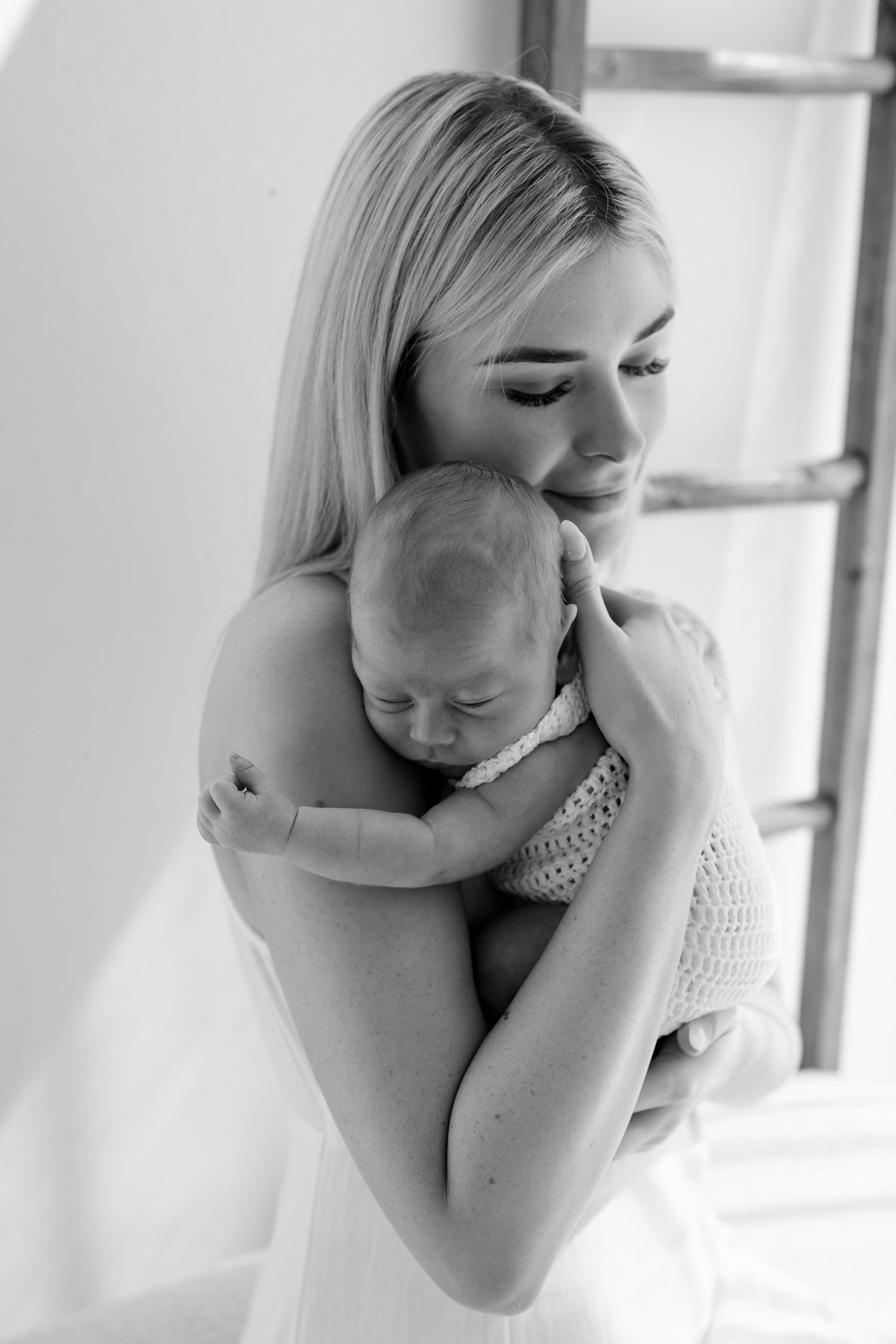 A tender black and white portrait of a mother holding and gently embracing her sleeping newborn baby by a window.