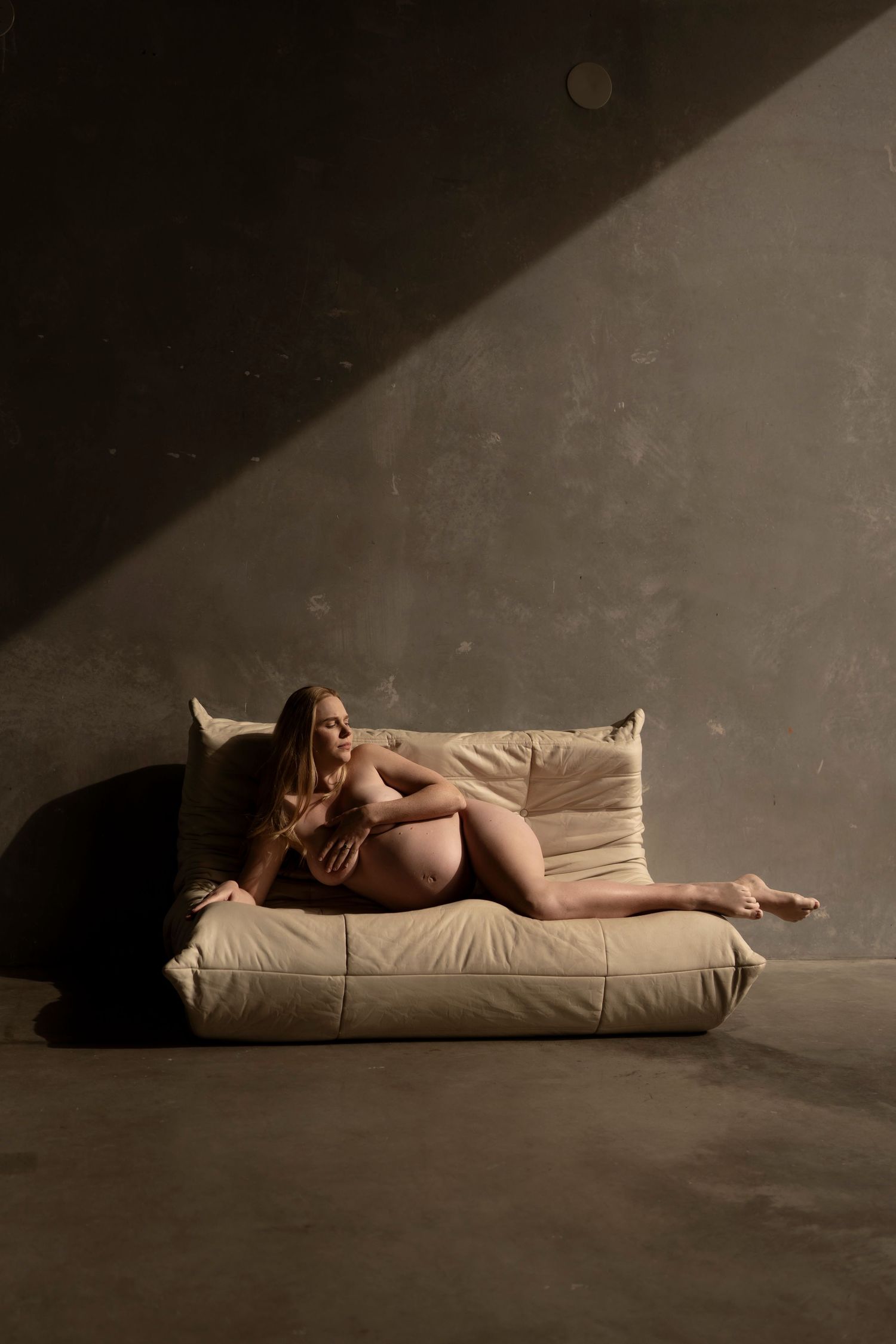 Pregnant woman reclining gracefully on a beige leather sofa in a moody, dimly lit concrete room with dramatic shadows.