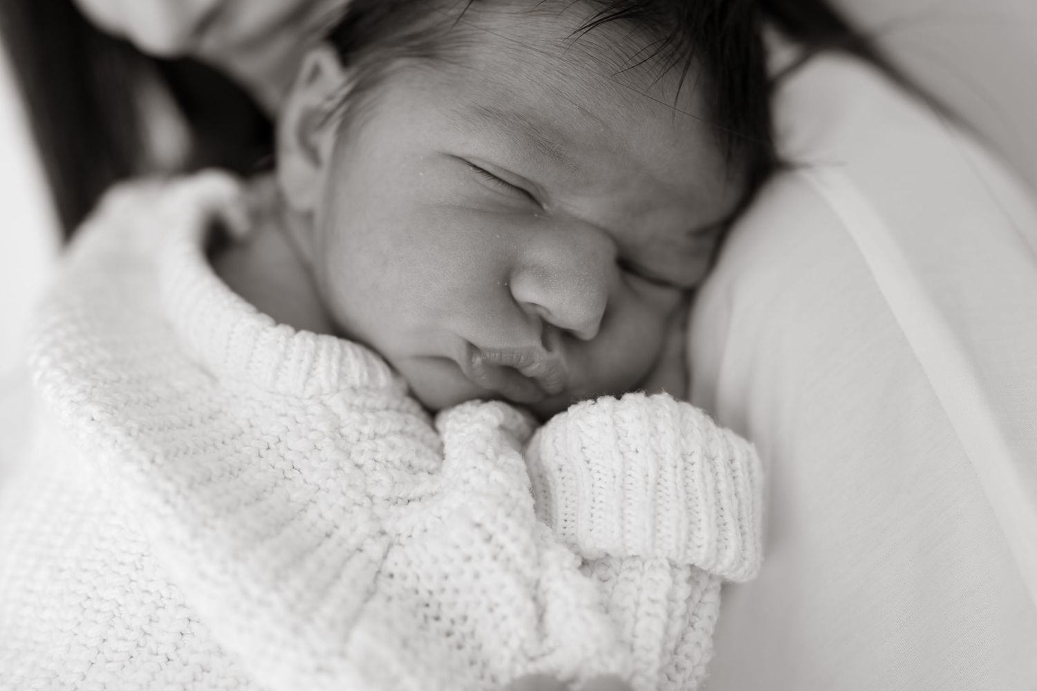A peaceful sleeping newborn baby wrapped in a soft white knitted blanket in black and white photography.