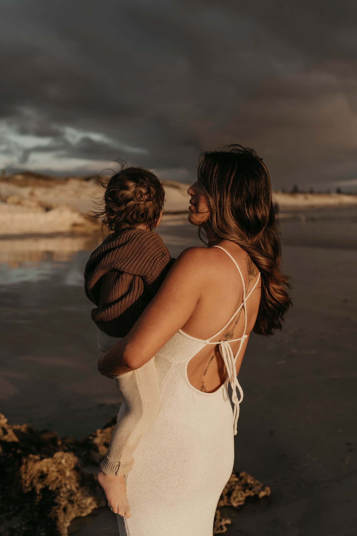 A silhouette of a woman in a white dress holding a child on a beach at dusk.