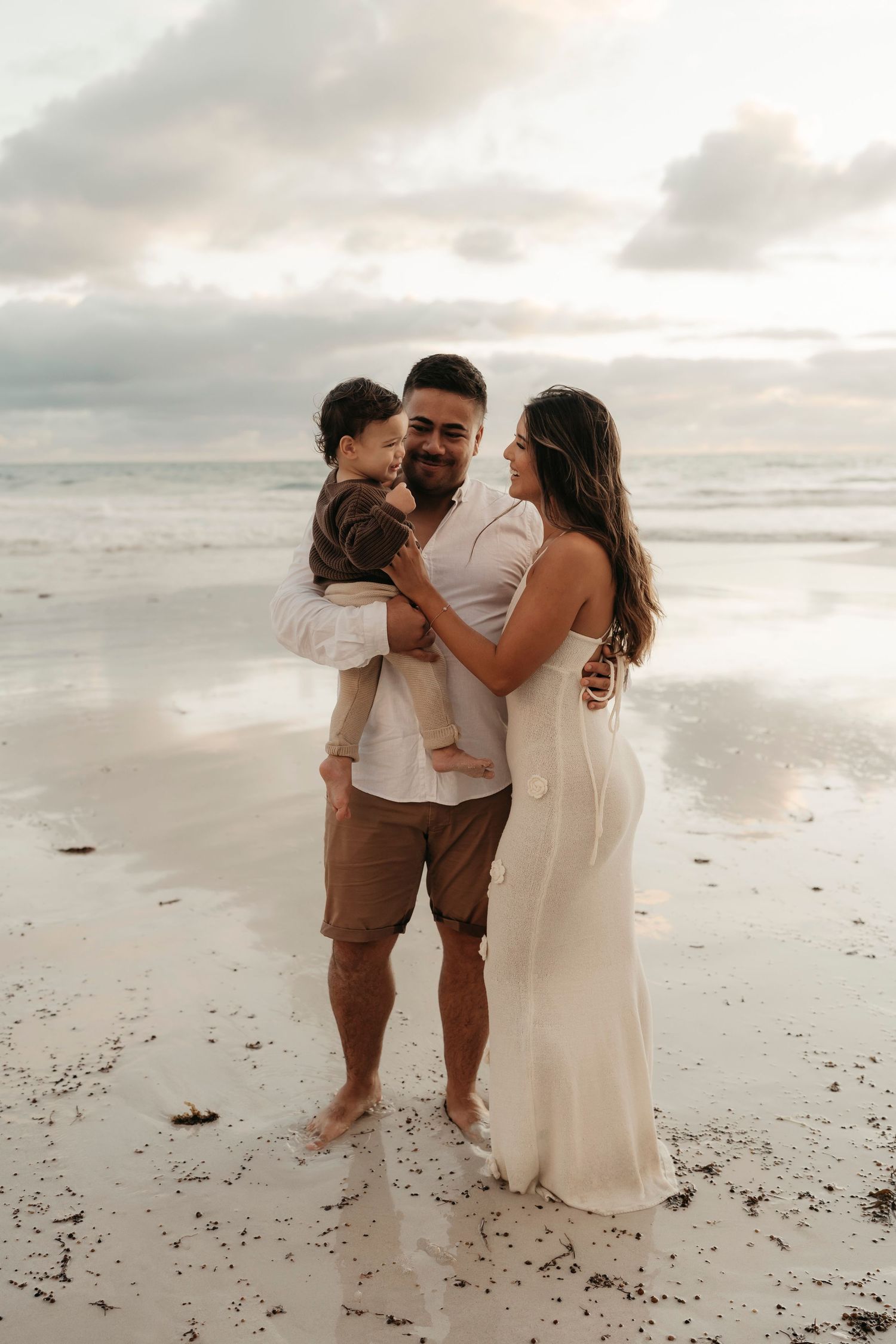 A family shares an intimate moment together on a cloudy beach, dressed in white and earth tones.