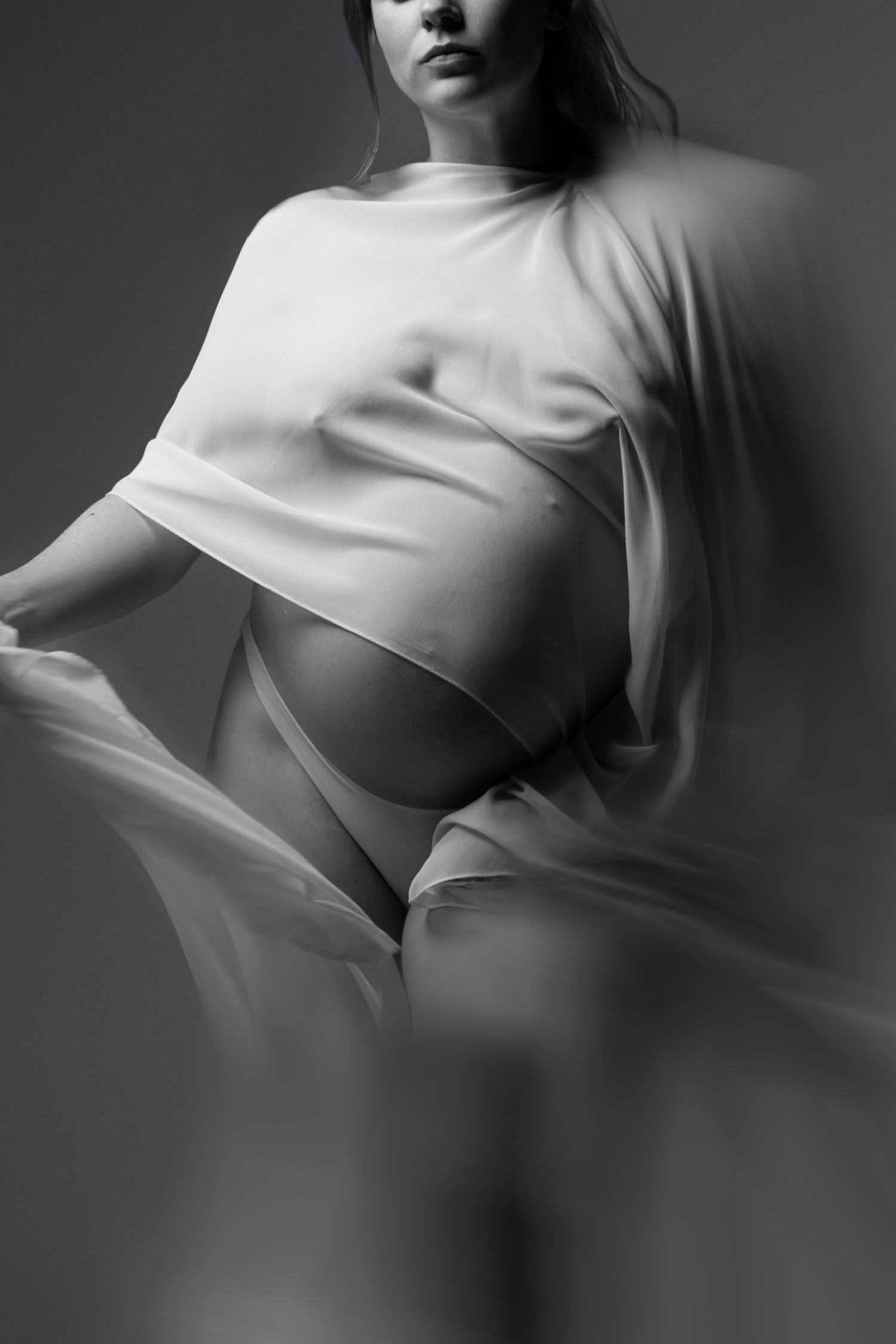 Black and white maternity photo of a pregnant woman draped in flowing white fabric, showcasing her baby bump.