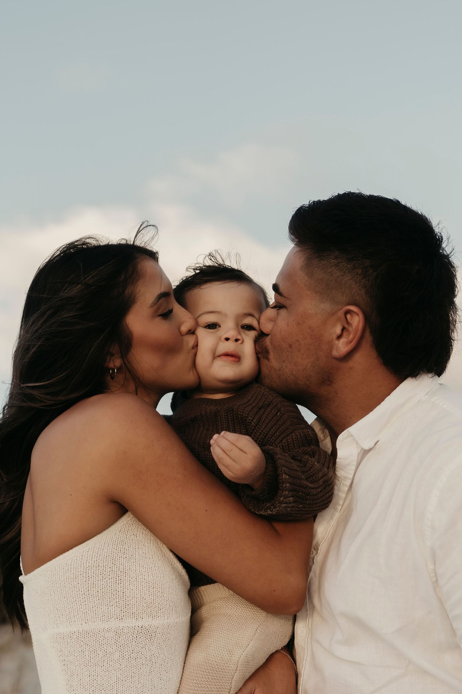 Sweet family moment captured outdoors with parents kissing their small child's cheeks against a soft sky background.