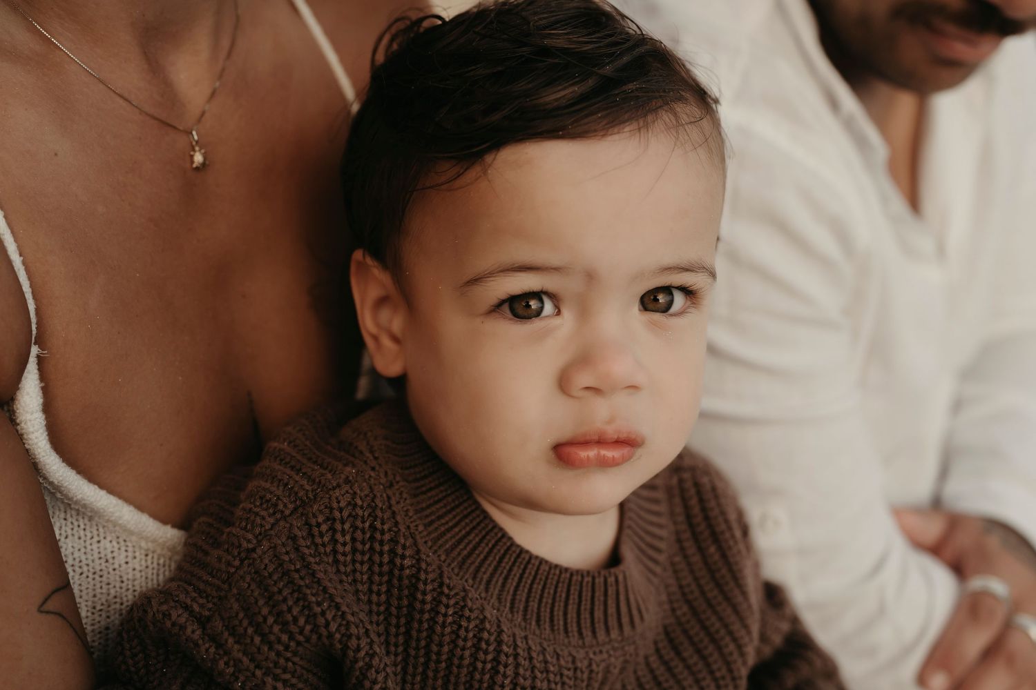 A small child wearing a cozy brown knit sweater sits between two adults in white clothing.