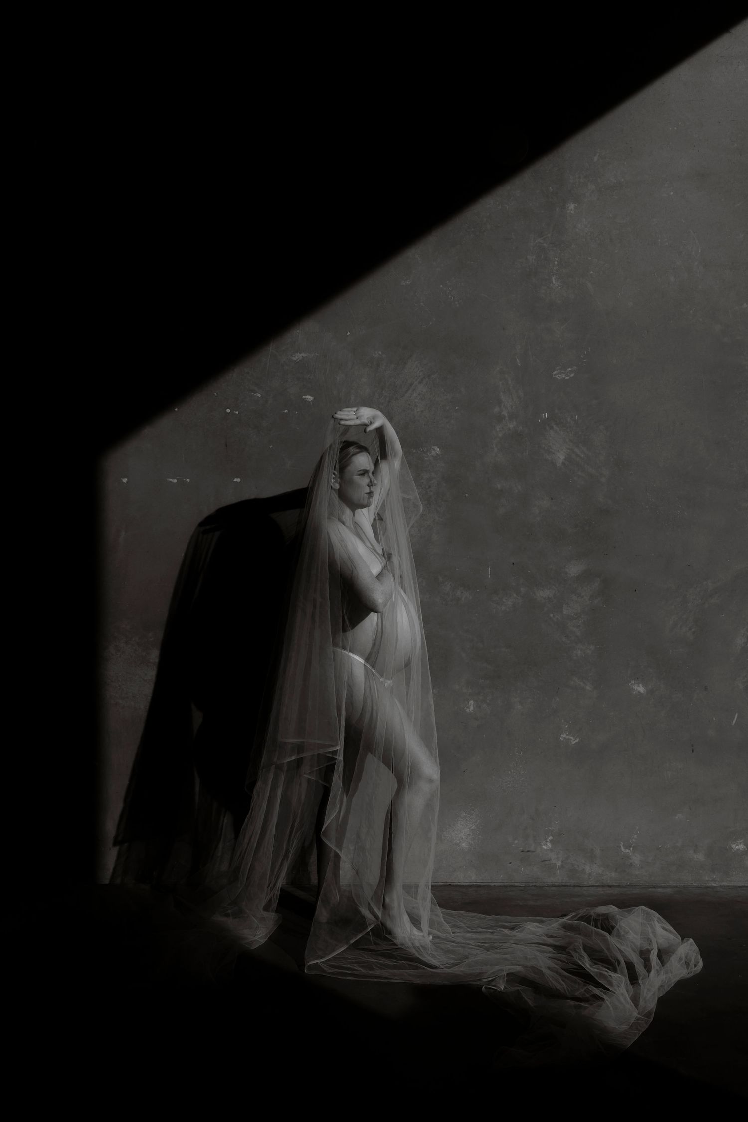 A woman in sheer white lingerie and veil poses dramatically in moody black and white boudoir photography.