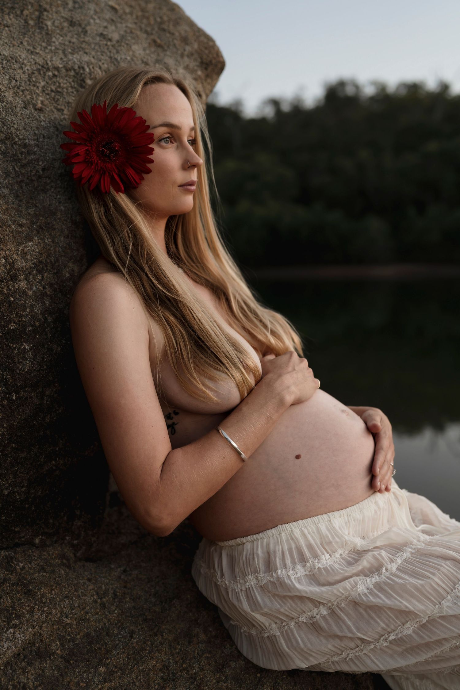 Maternity portrait featuring a red flower accent, white dress, and natural rock backdrop near a peaceful lake.