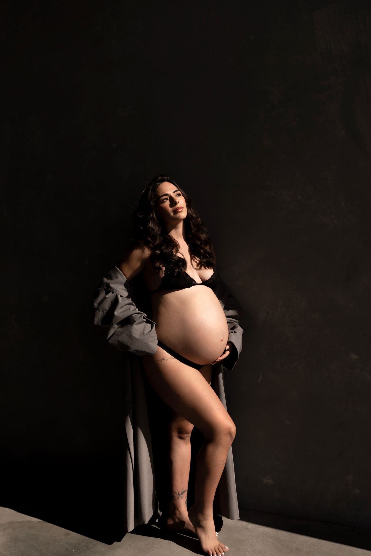 Pregnant woman in black lingerie and open gray robe posing against dark studio background, showcasing baby bump.