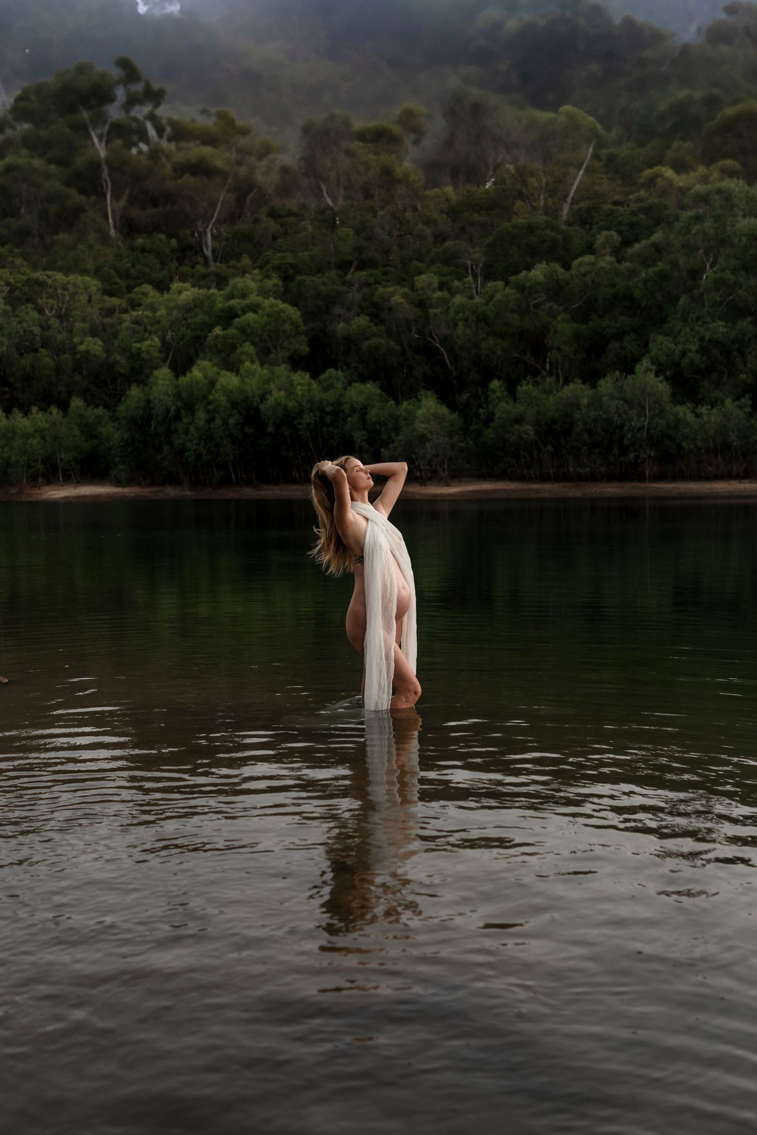 Ethereal figure in flowing white dress stands in calm lake water surrounded by misty forest at dusk.
