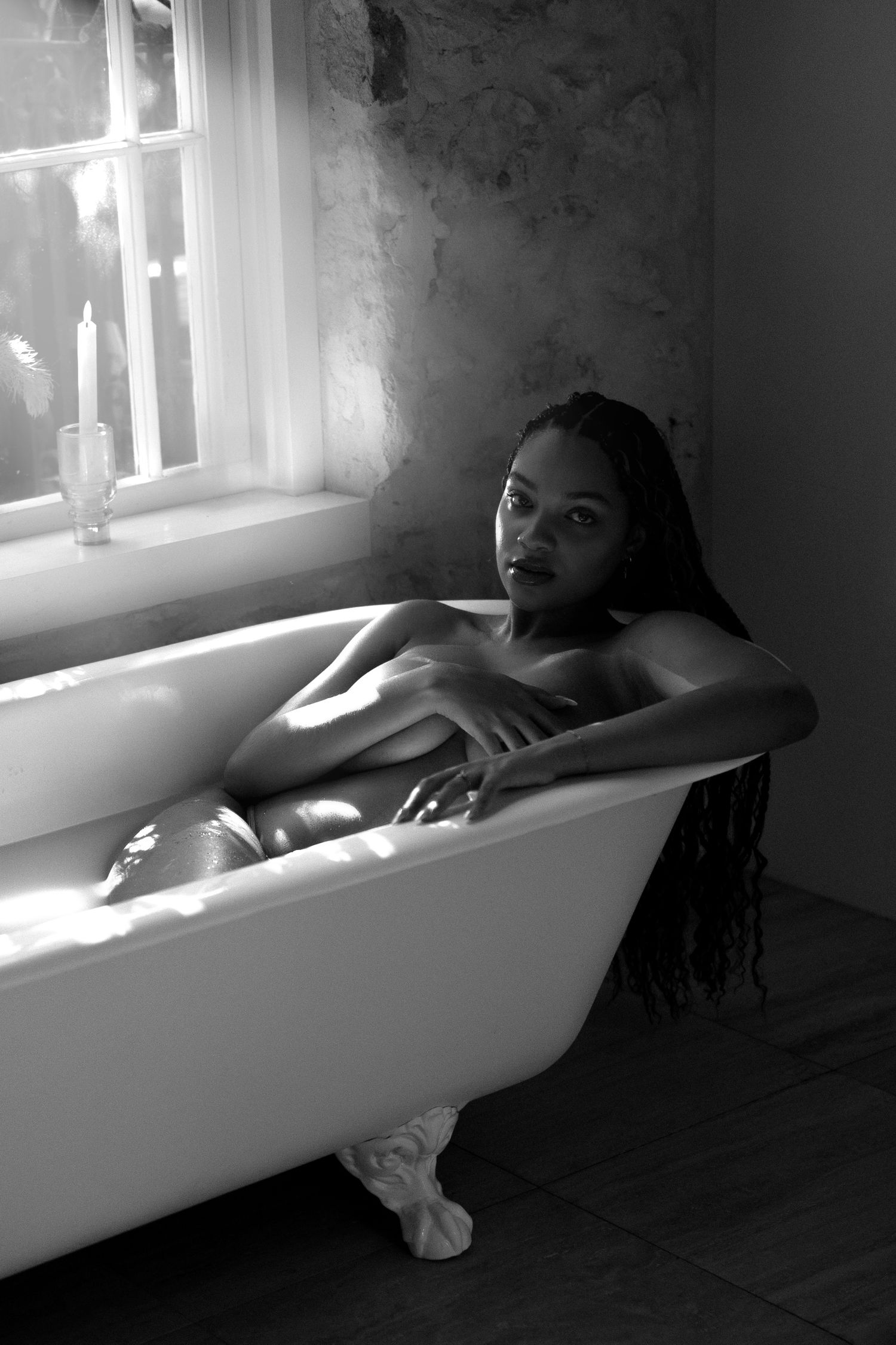 Black and white photo of a woman relaxing in a vintage clawfoot bathtub near a window with candles.
