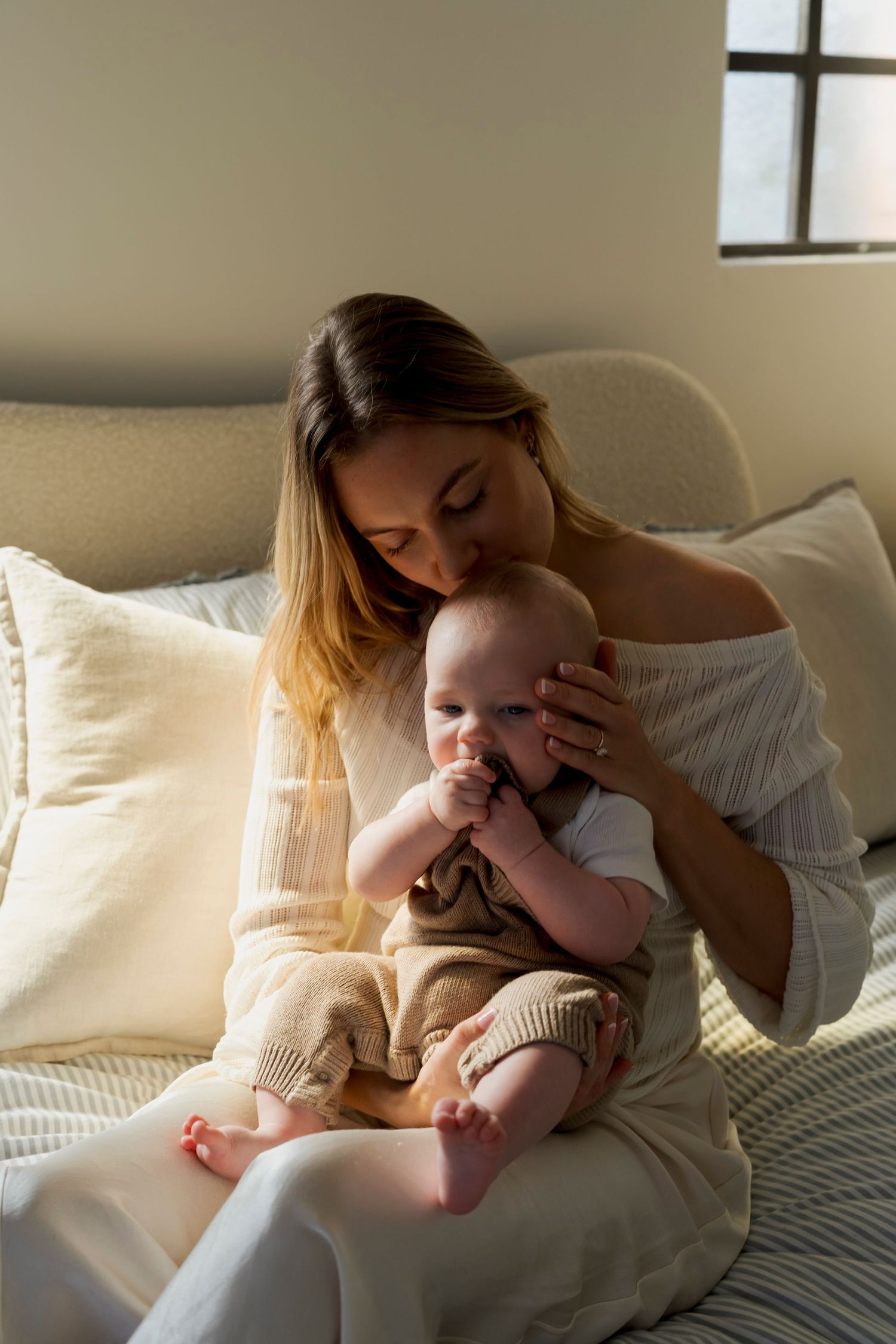 A tender moment at home as a parent in white comforts their baby on a cozy bed with soft natural lighting.