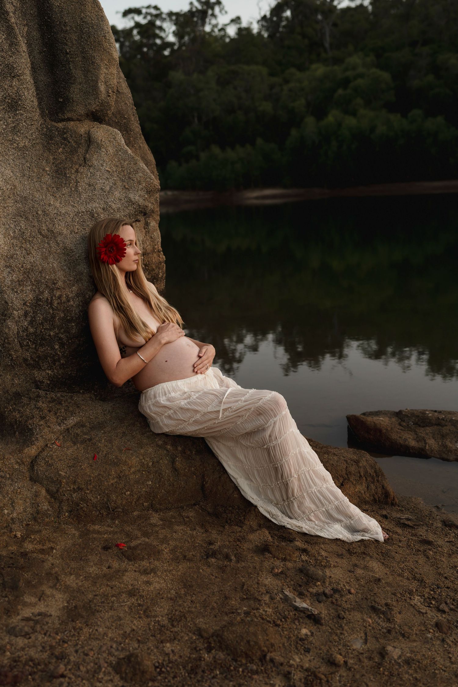 Artistic maternity photo of a woman in white dress reclining against rocks by a serene lake at dusk.