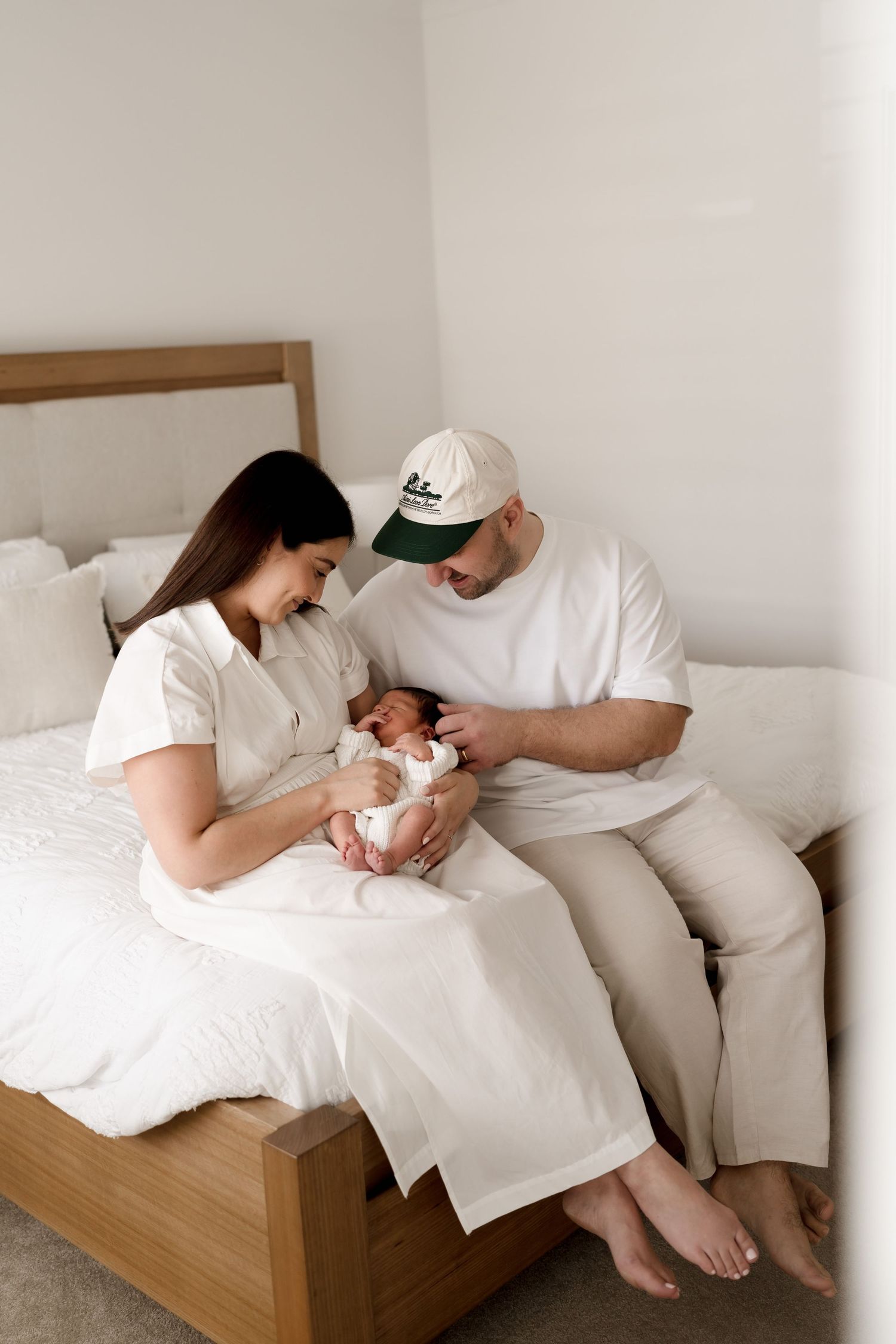 Family enjoys tender moment together on bed with newborn baby in soft white minimalist bedroom setting.
