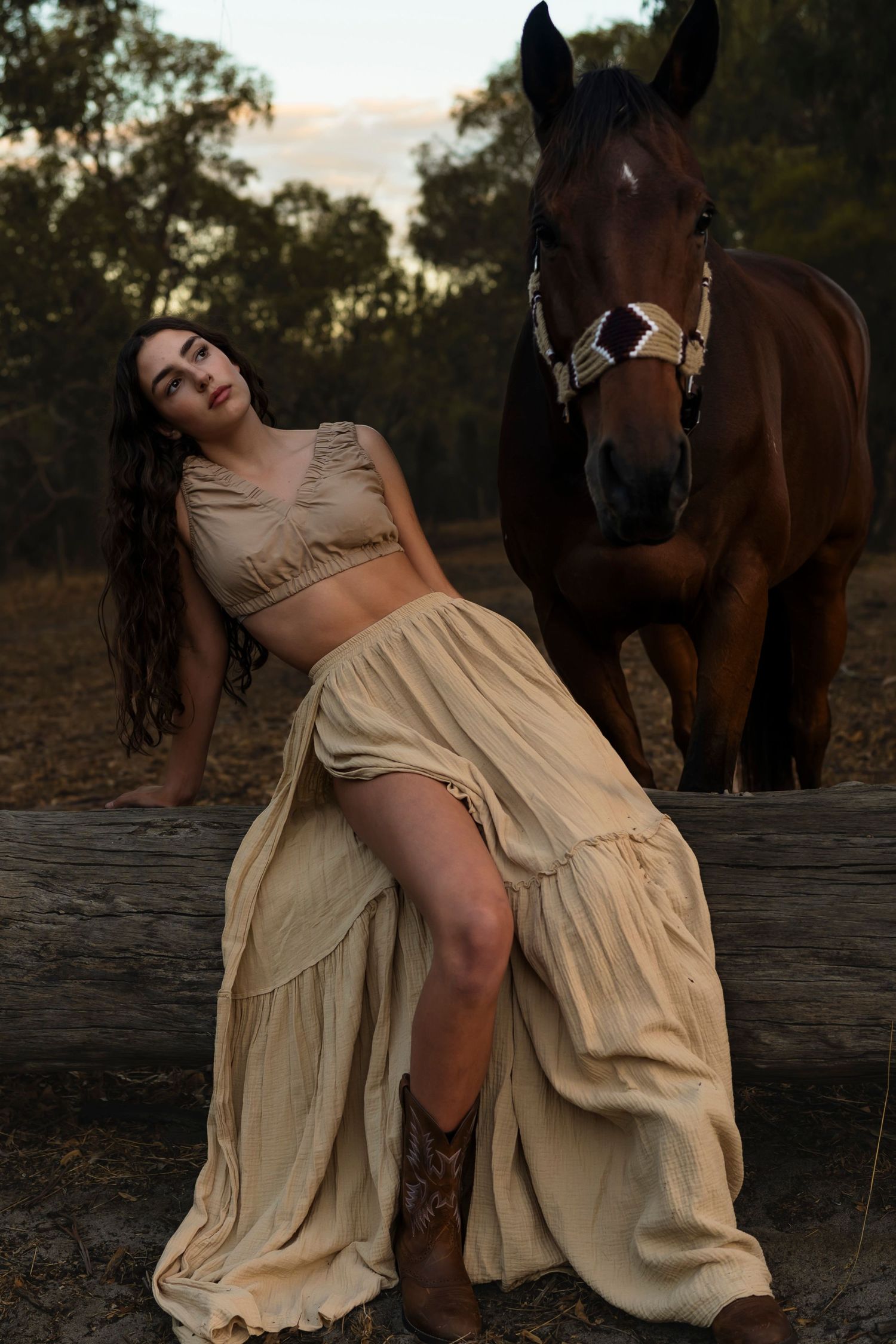 A brown horse stands beside a crop top and flowy skirt at sunset in a rustic outdoor setting.