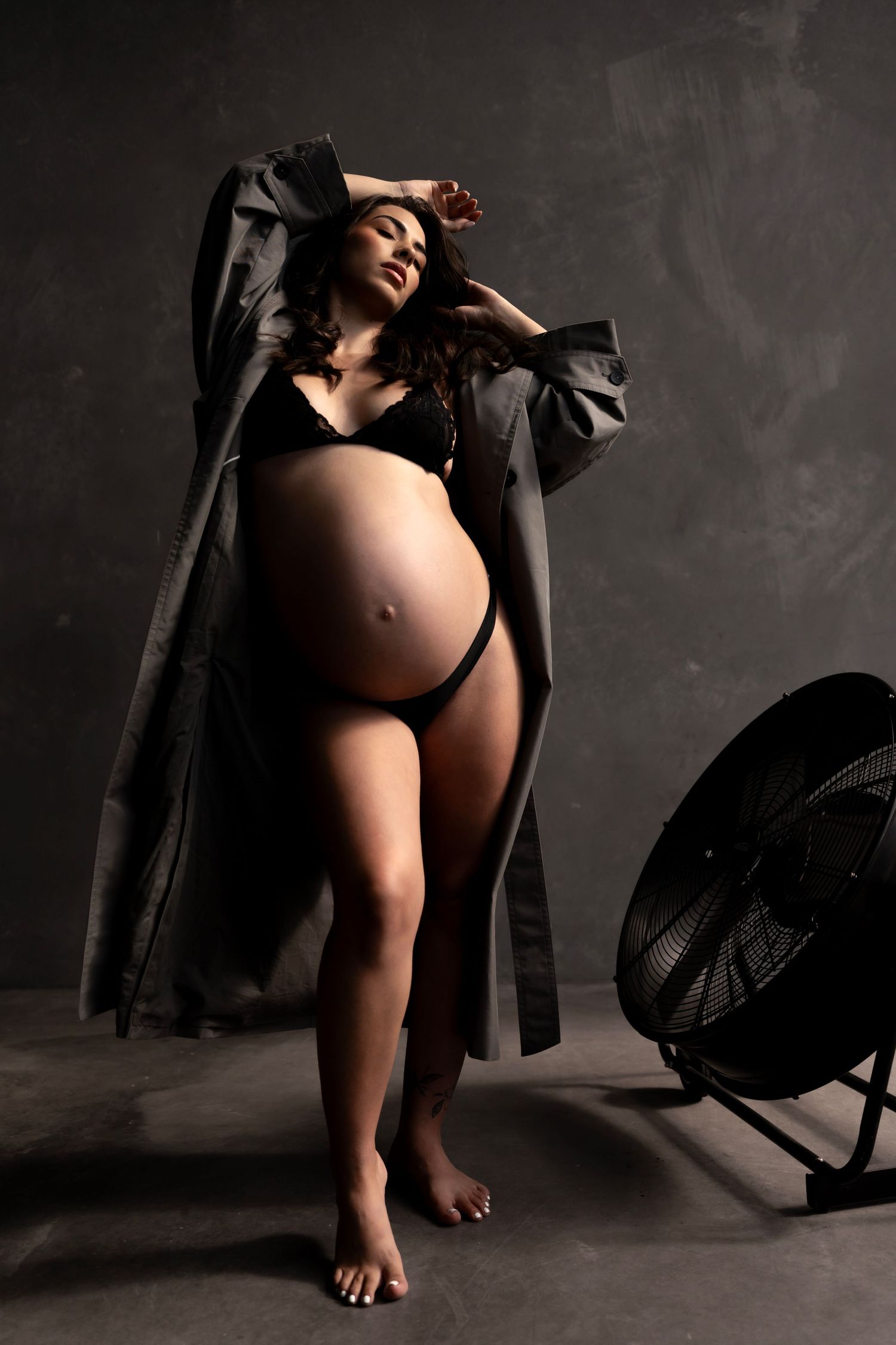 Pregnant woman in black lingerie and gray coat posing confidently in a dark studio with a floor fan nearby.