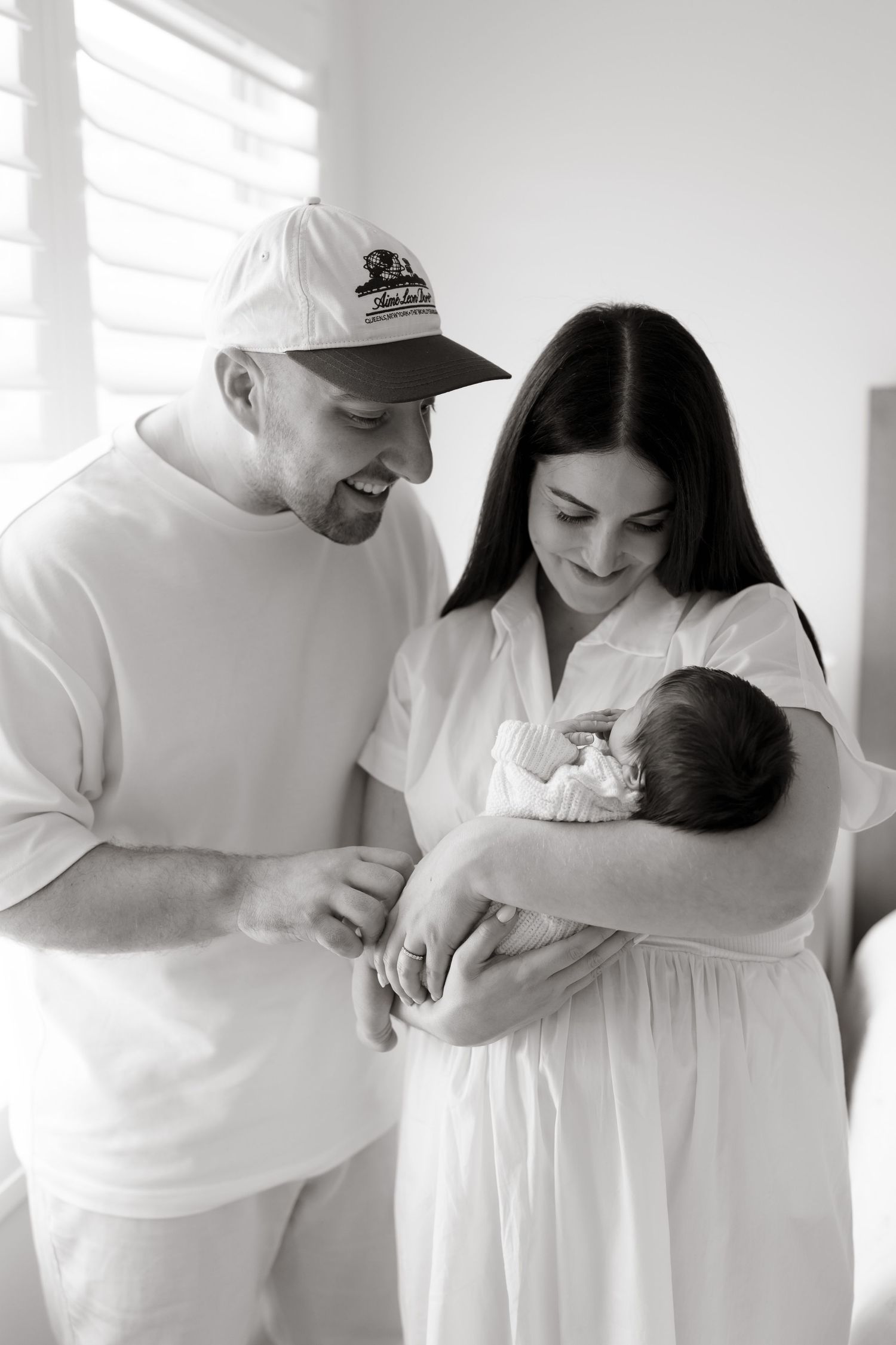 A serene black and white portrait of parents in white clothing admiring their newborn baby. Meg Rose Photos Perth WA.