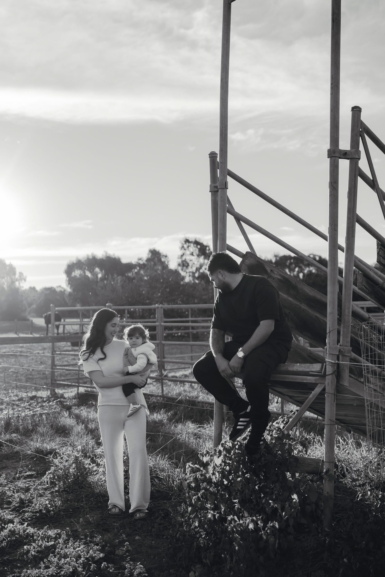 Black and white family photo of a woman holding a baby and a man sitting on rustic wooden steps in a rural farm setting.