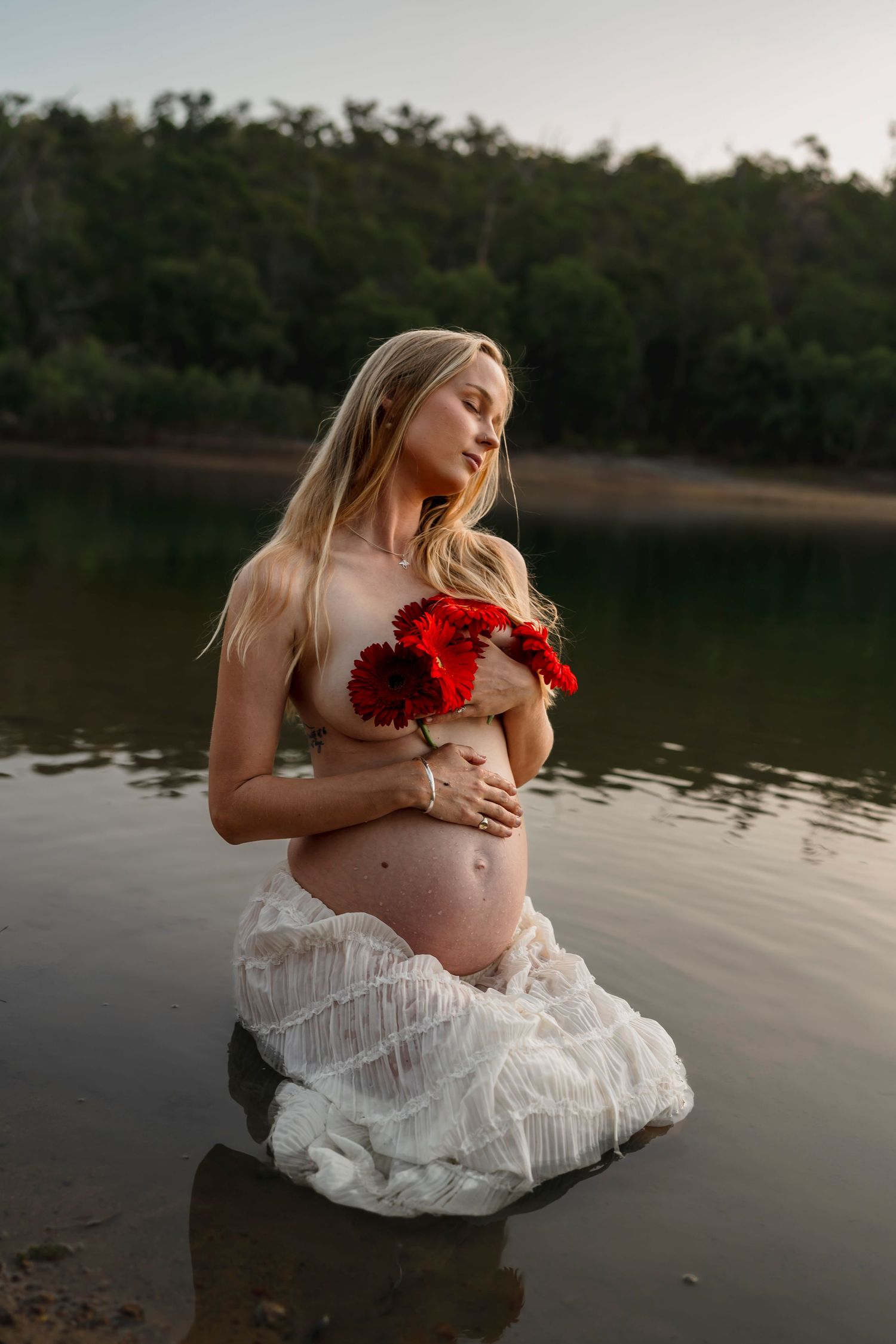 Artistic maternity photo at a lake with a white lace dress and red roses at sunset.