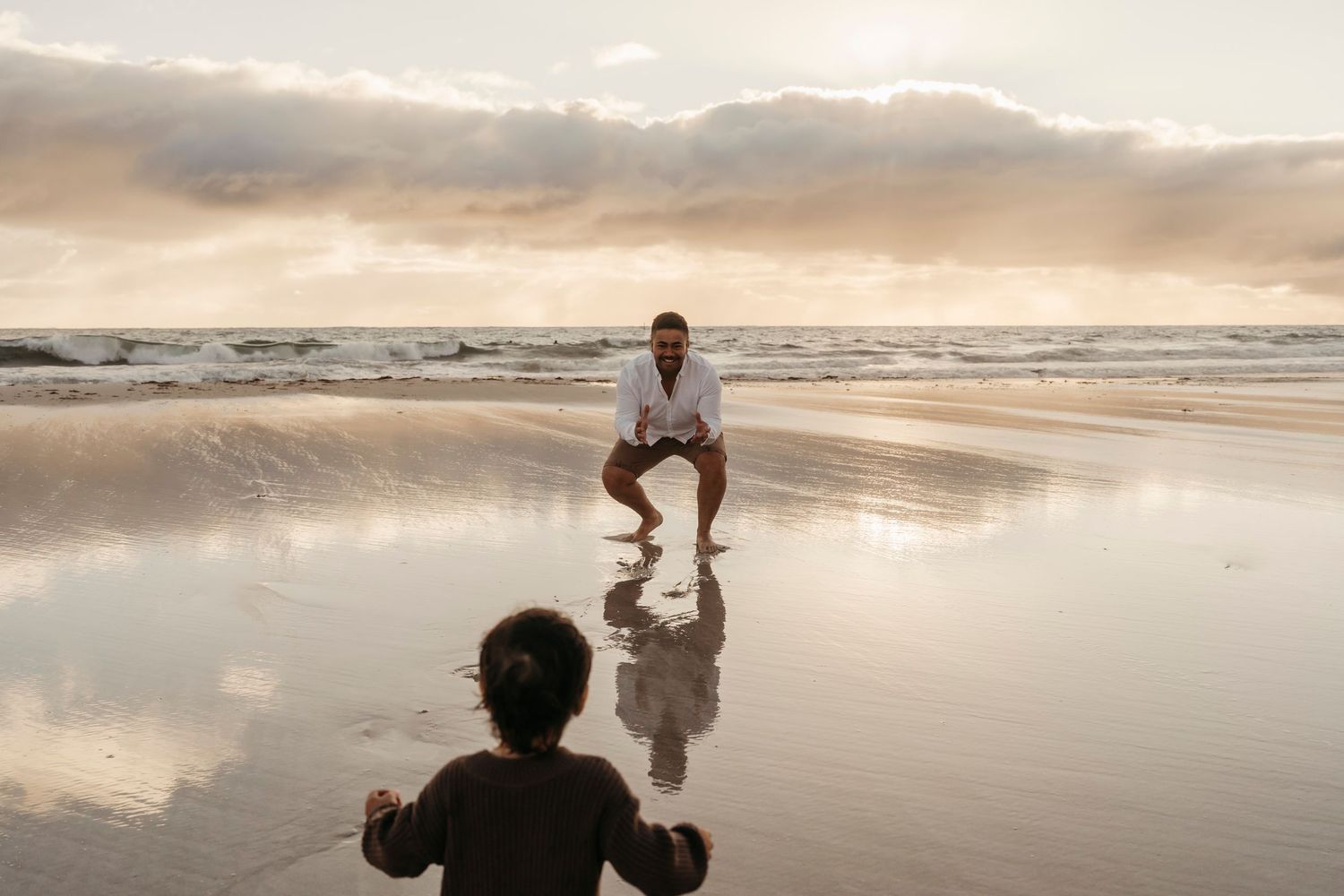 Two people on a reflective beach at sunset with waves and clouds in the background.