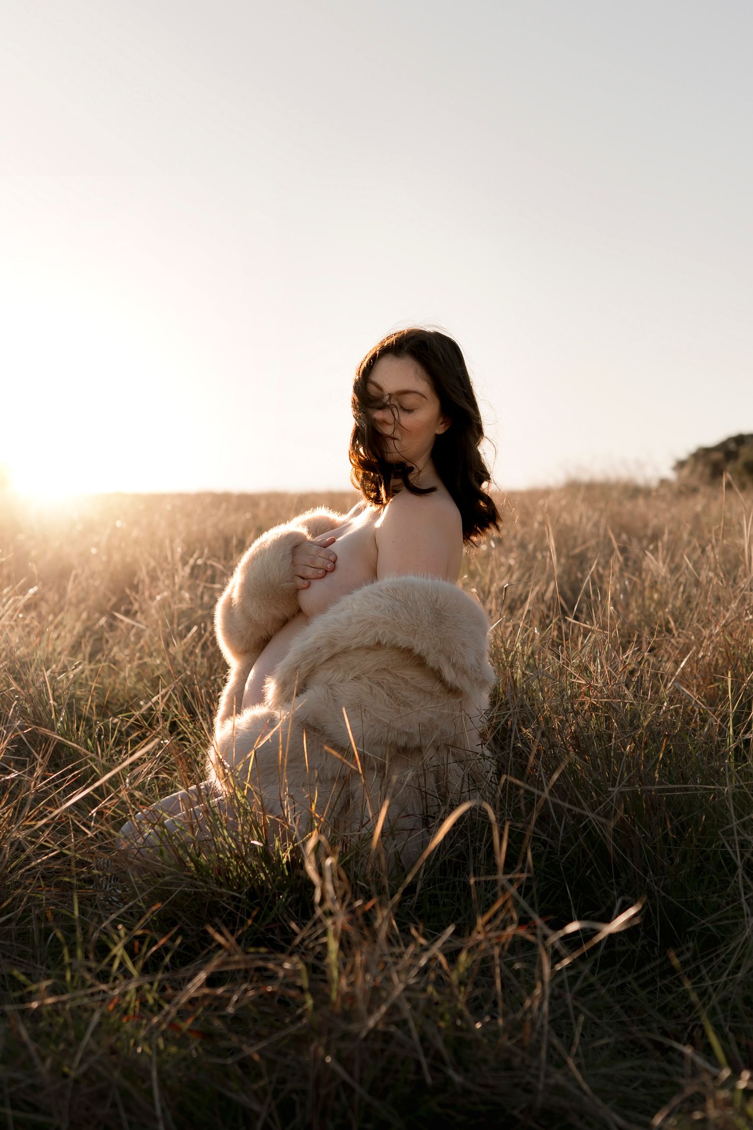 Woman in fluffy coat sitting in golden grass field at sunset.