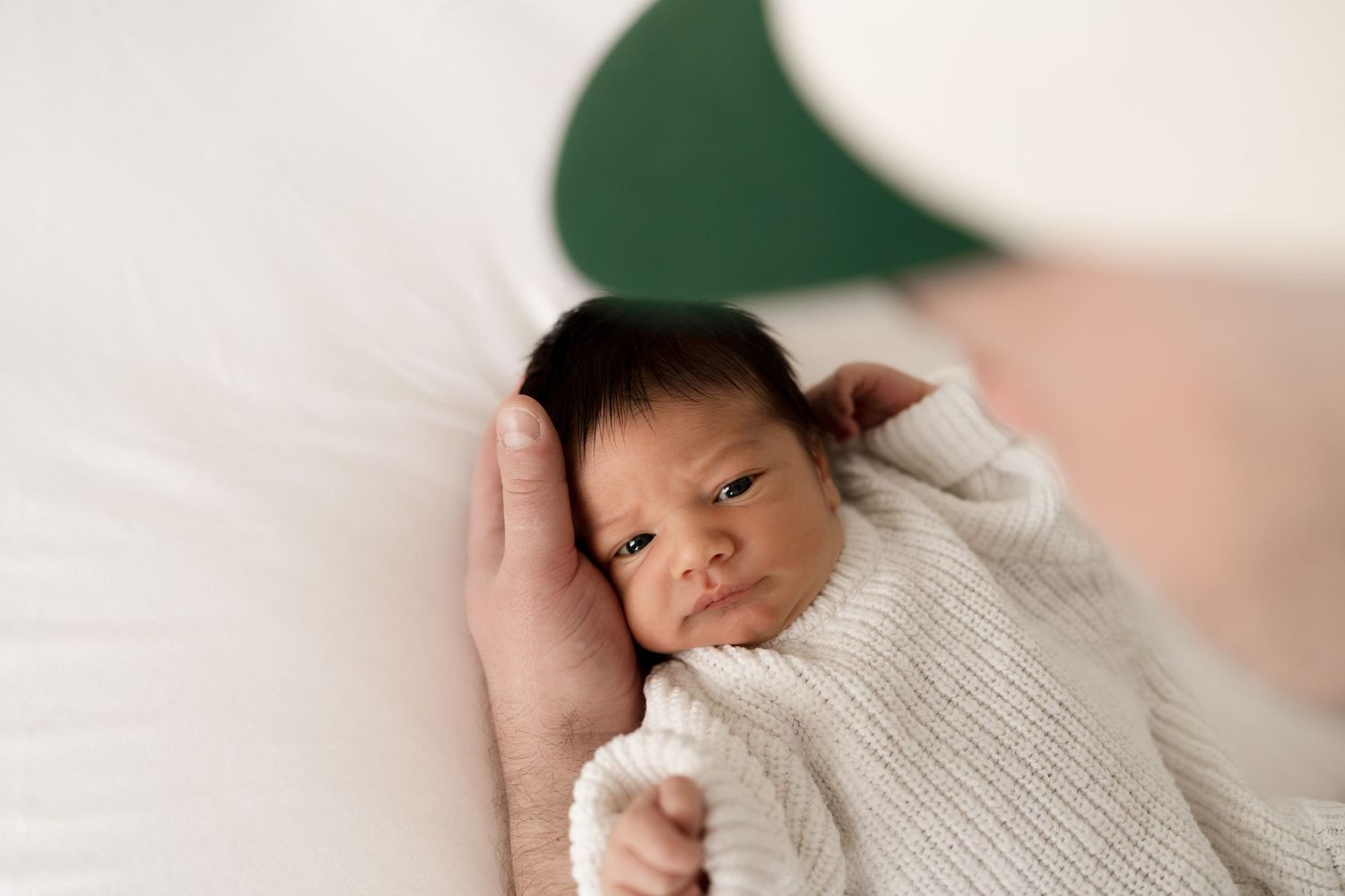 Newborn swaddled in white knit sweater being cradled against green background. Meg Rose Photos Perth WA.