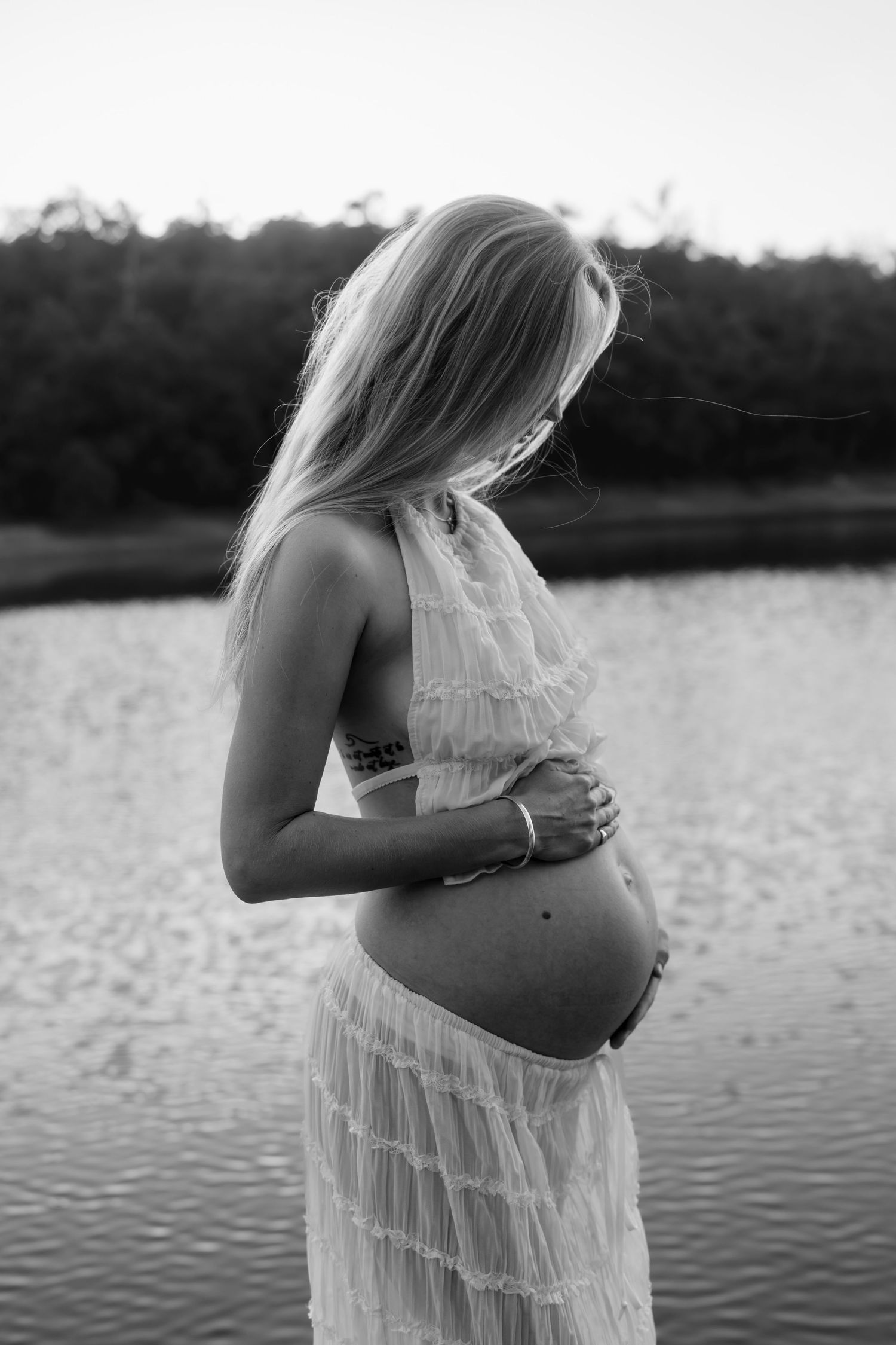 Artistic maternity photo of an expecting mother in a white dress standing by a serene lake at sunset.
