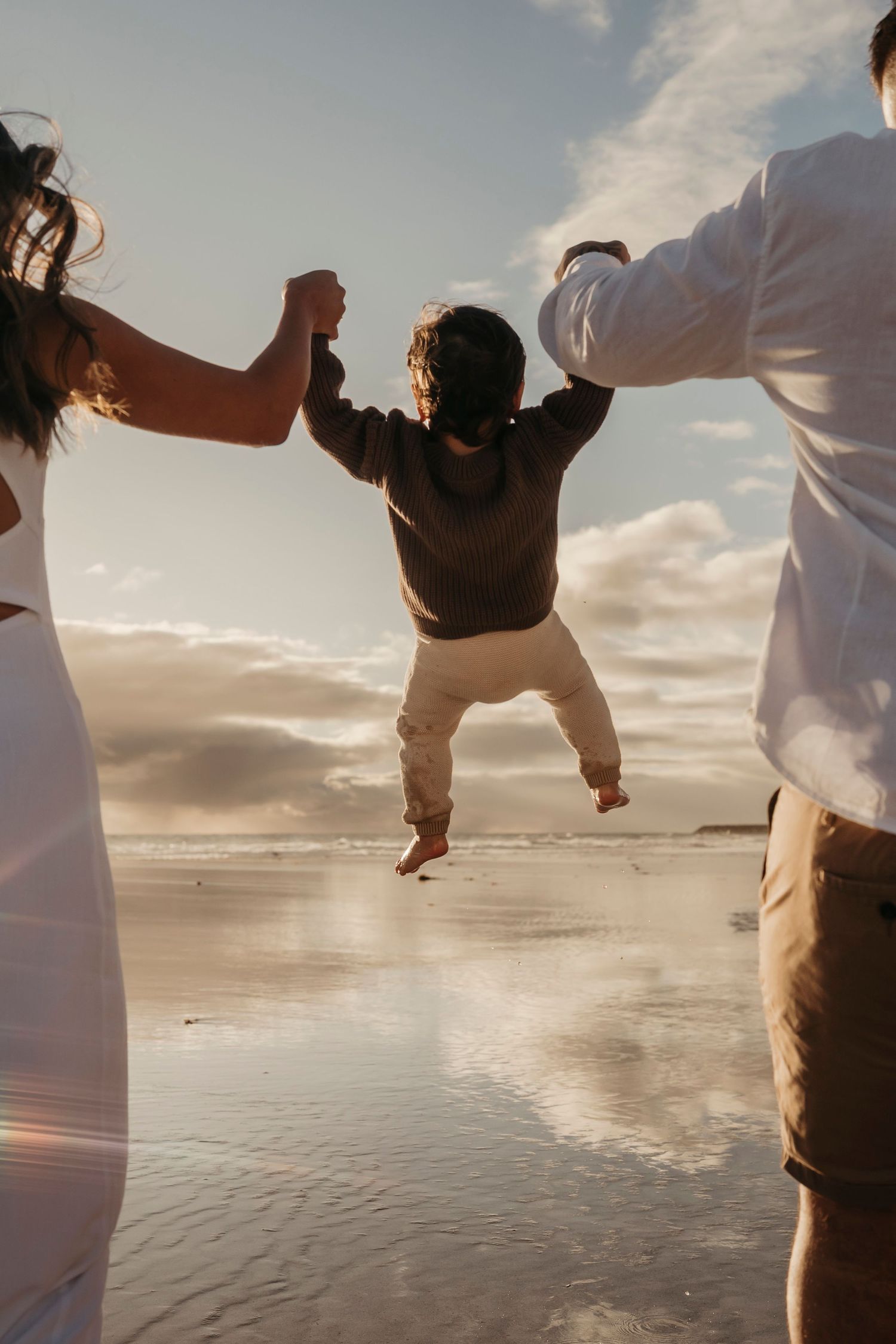 Small child being lifted up by adults while playing on a sunny beach at sunset with ocean waves in the background.