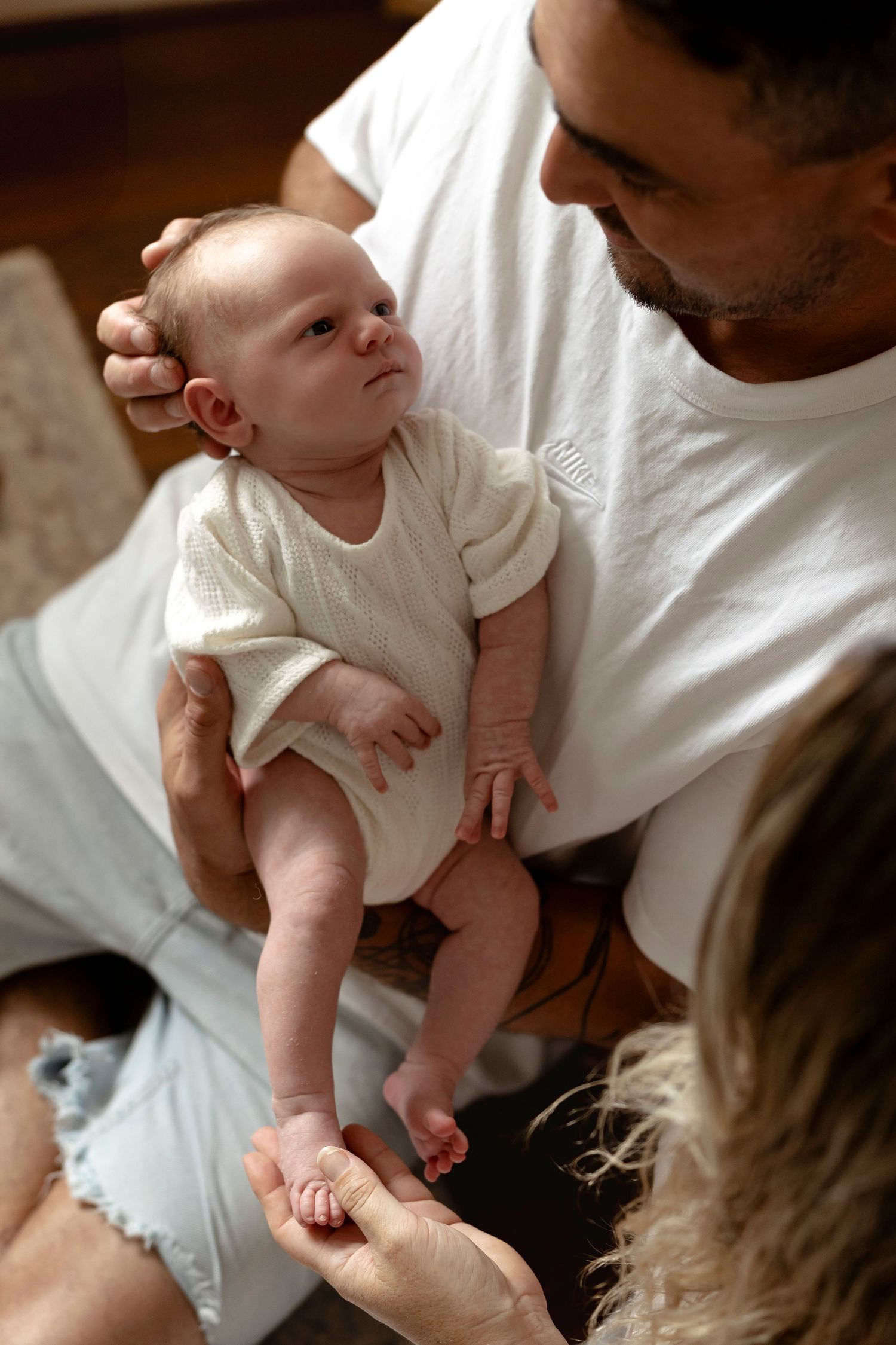 A father holds his newborn baby in a white onesie while a mother gently touches the infant's feet indoors.