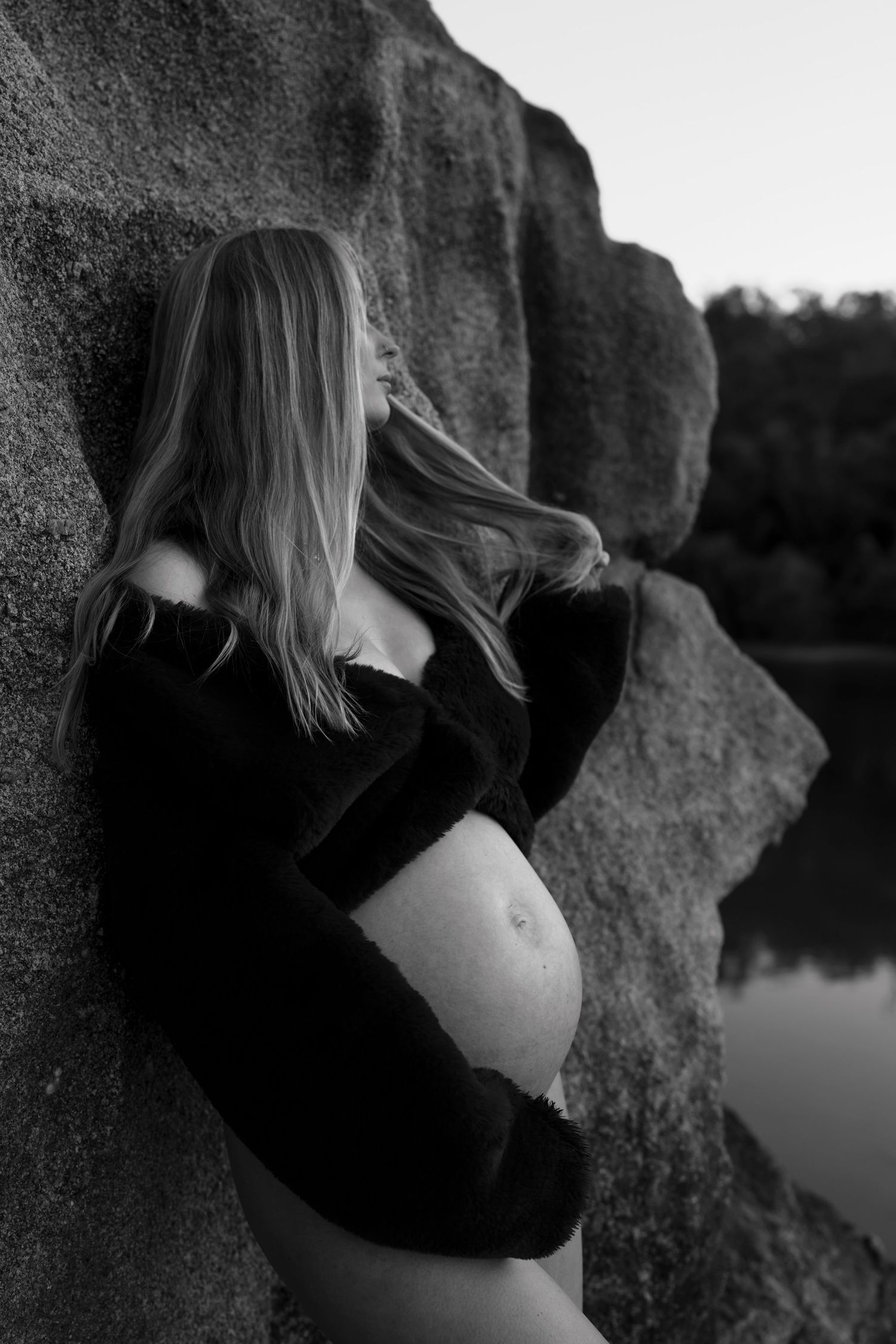 Artistic maternity photo of woman in black dress against cliffside by water.