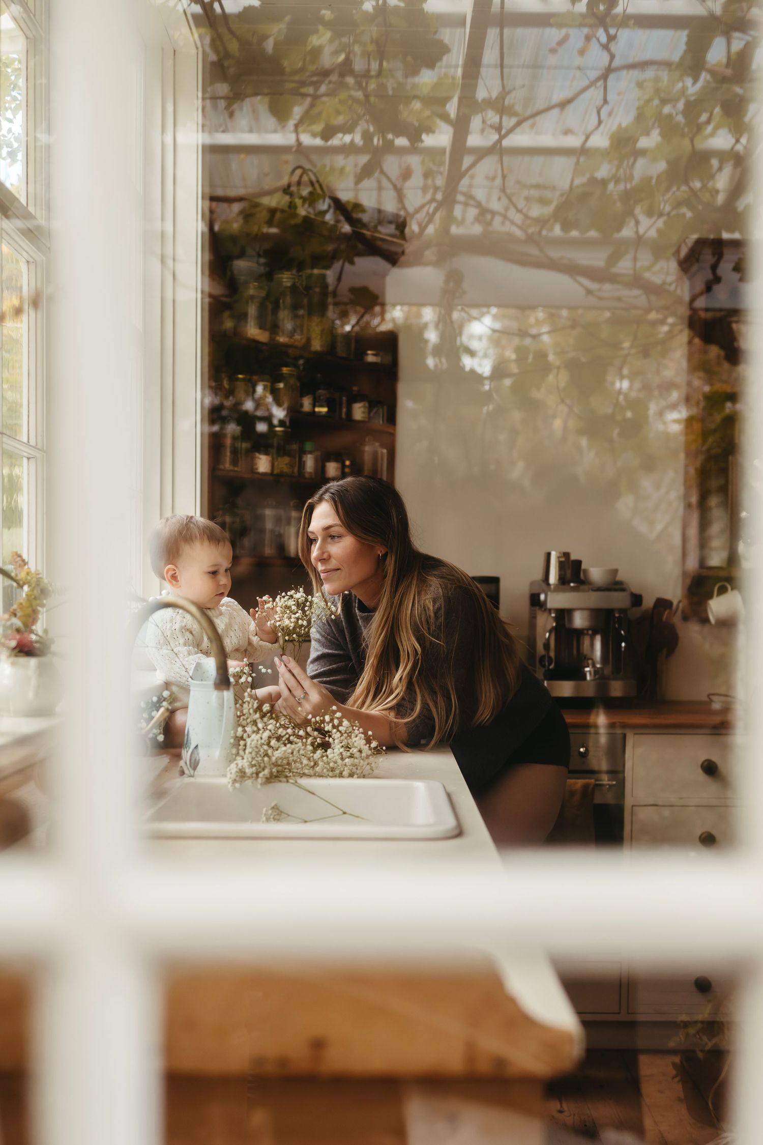 A mother smiles at her baby near a kitchen sink, surrounded by plants and natural light.