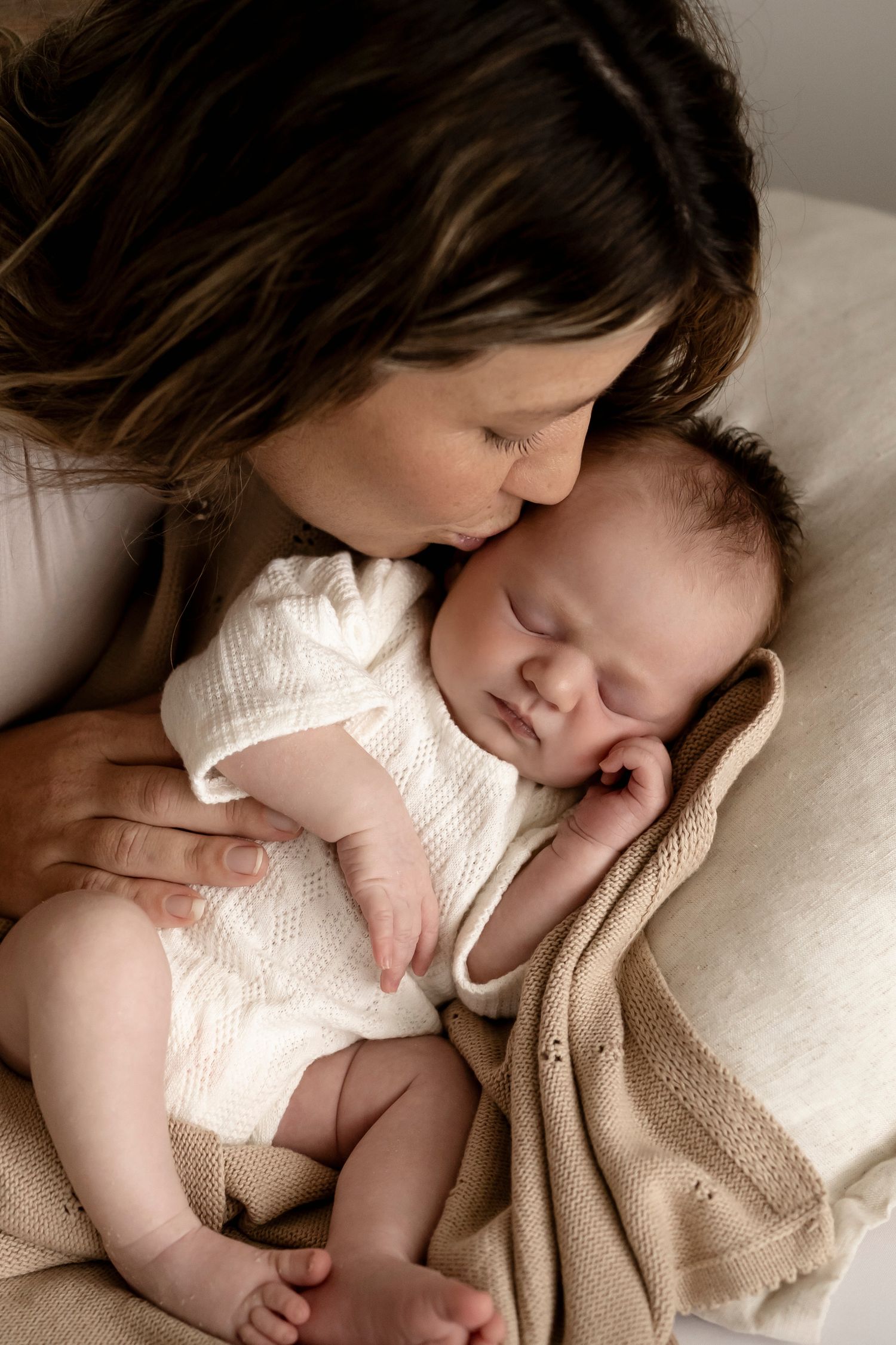 A mother gently kisses her sleeping newborn baby dressed in white, wrapped in a soft beige knit blanket.