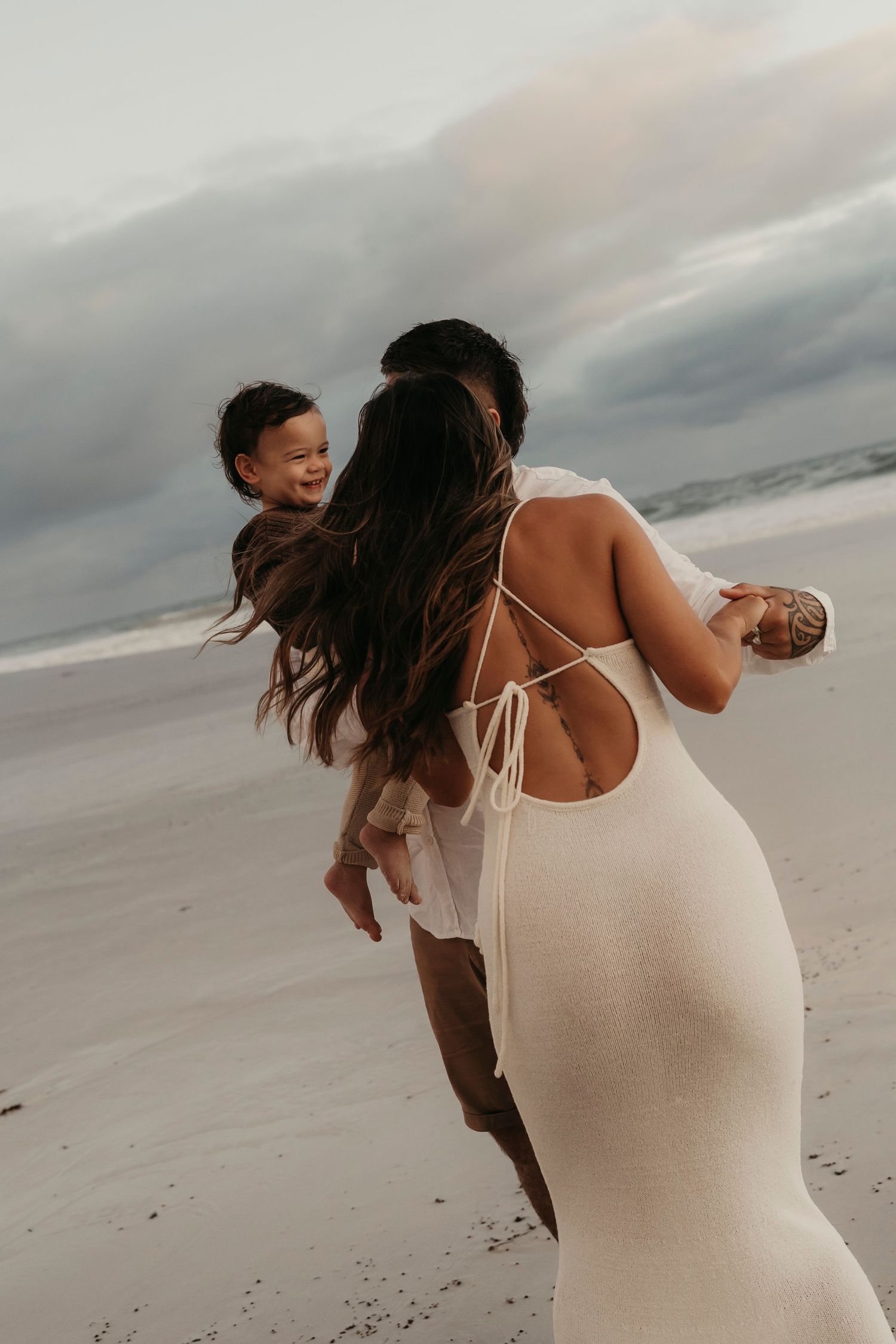 A parent in a white dress spins playfully with a small child on a cloudy beach.