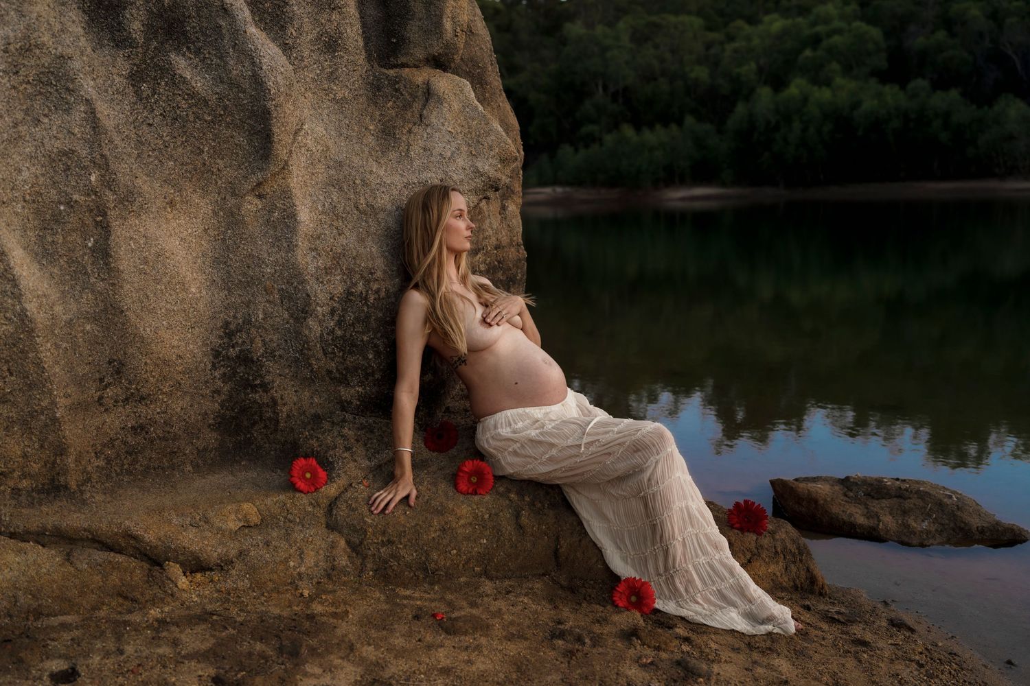 Figure in white lace dress poses on rocky outcrop by lake with red roses scattered around.