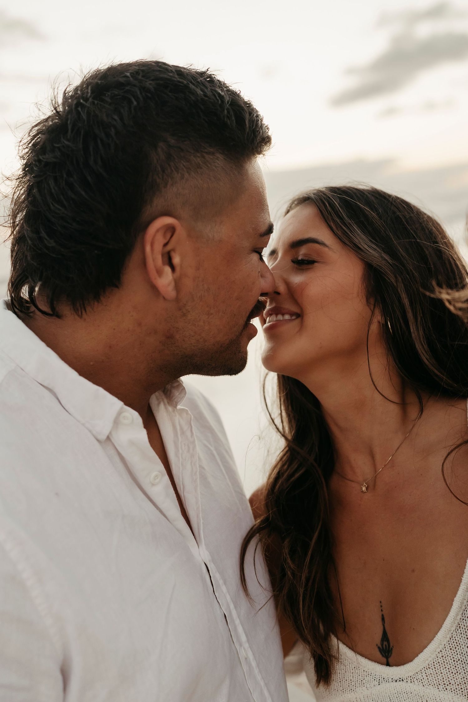 A romantic couple shares an intimate moment at sunset on the beach while wearing white attire.