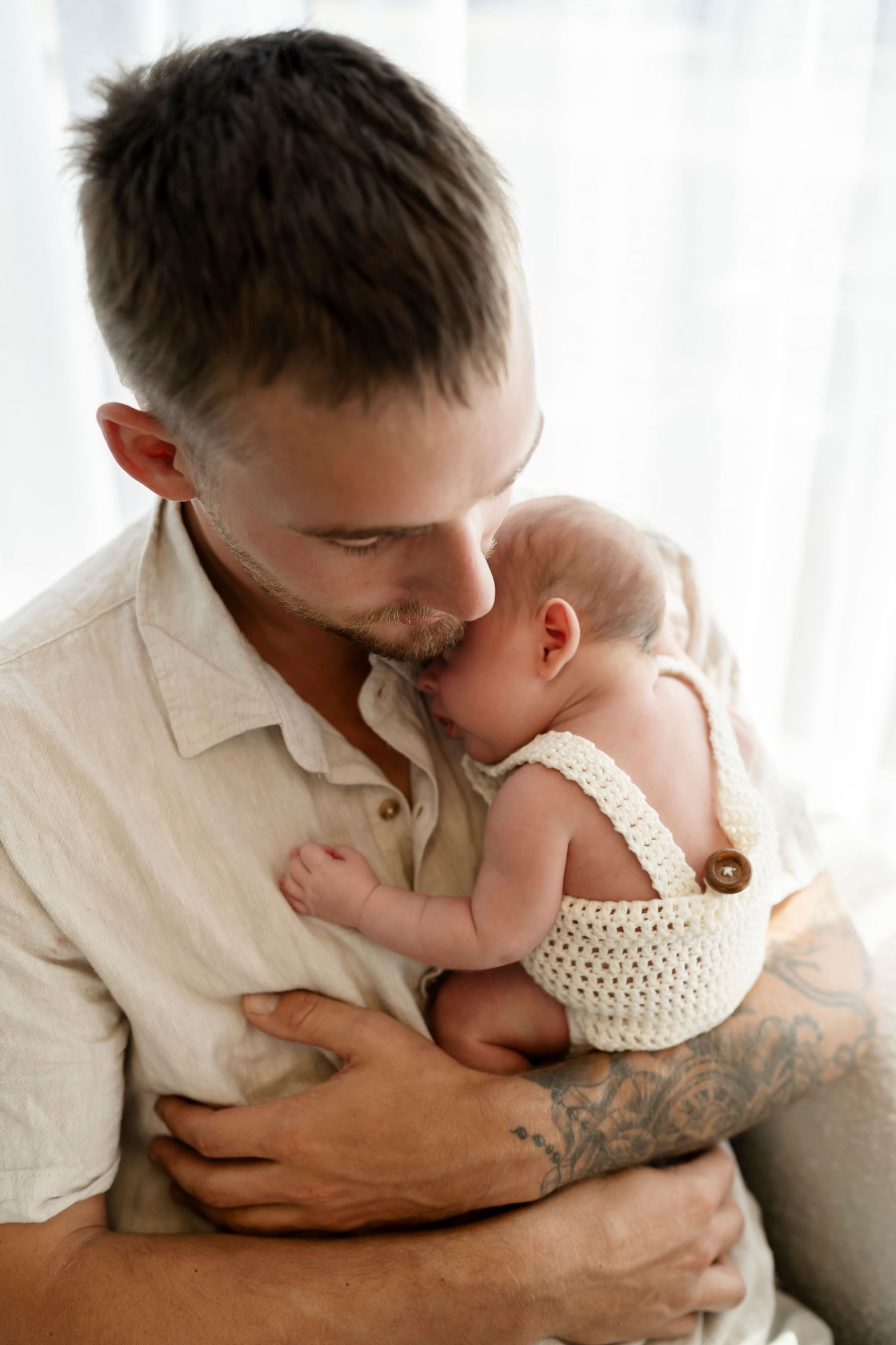 A loving parent tenderly holds and kisses their newborn baby wearing a crocheted outfit against a bright window backdrop.