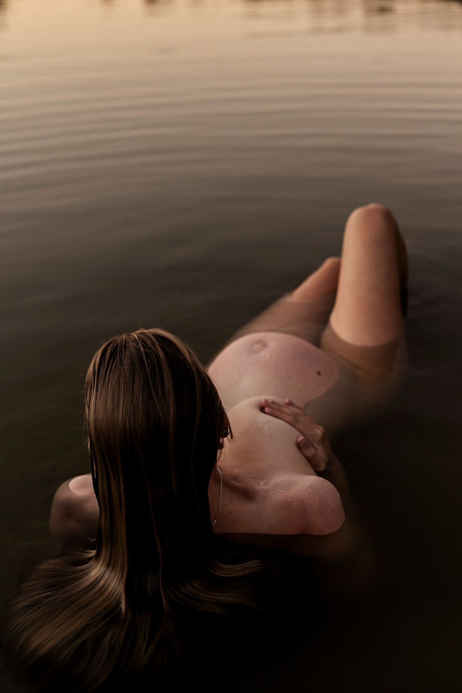 An artistic maternity photo captures a pregnant woman floating peacefully in dark, still waters at dusk.
