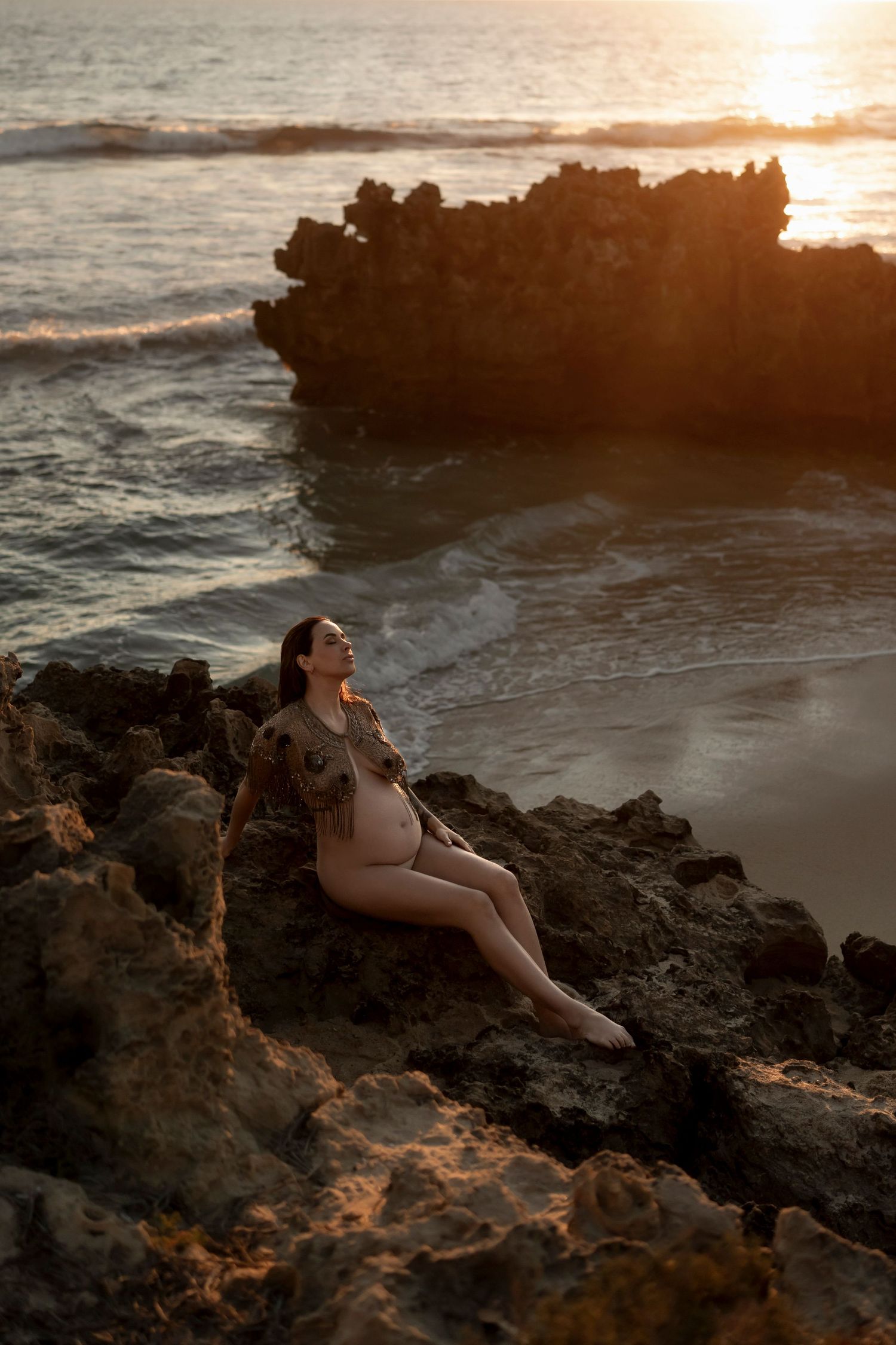 Pregnant woman in sheer bodysuit sitting on rocky shoreline at golden sunset with ocean waves in background.