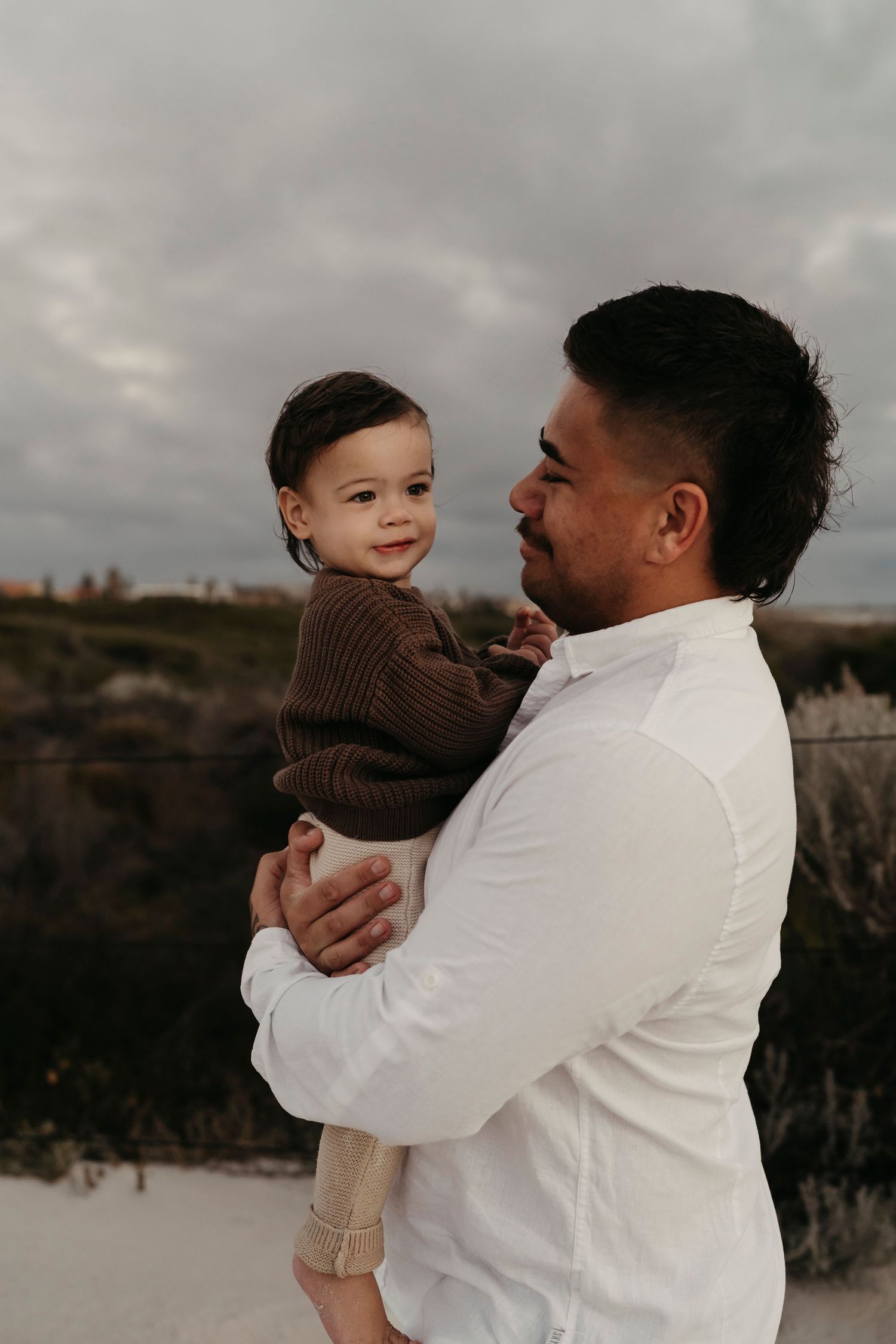 A young child in a brown sweater being held lovingly by someone in a white shirt against a moody sky.