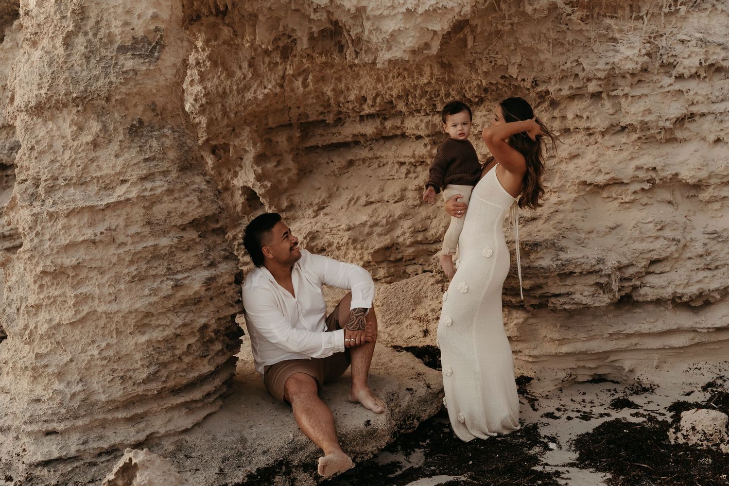 Two figures share a moment against a dramatic limestone cliff face on a rocky beach.