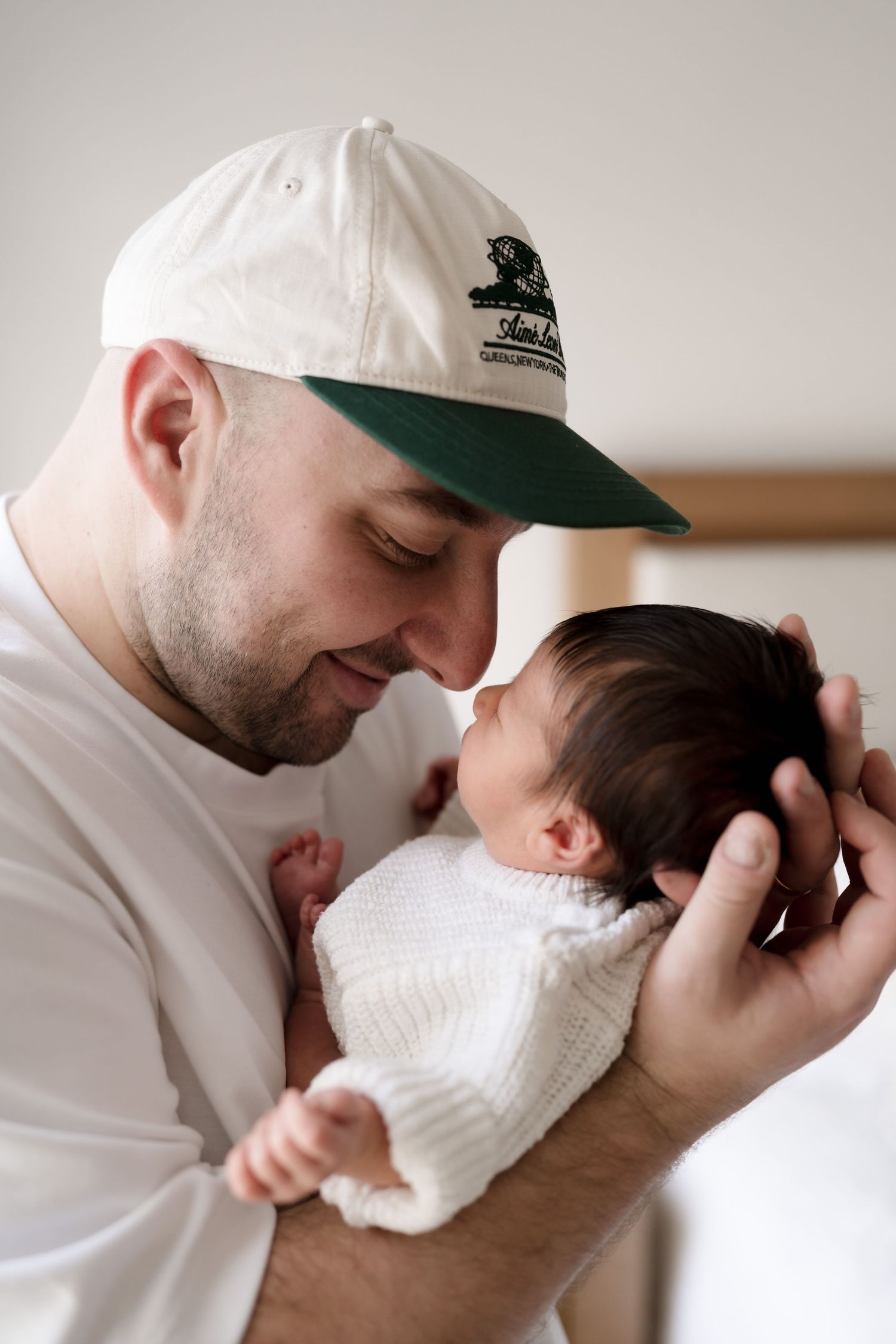 A tender moment showing an adult in a white baseball cap gently holding and cradling a newborn baby in white clothing.