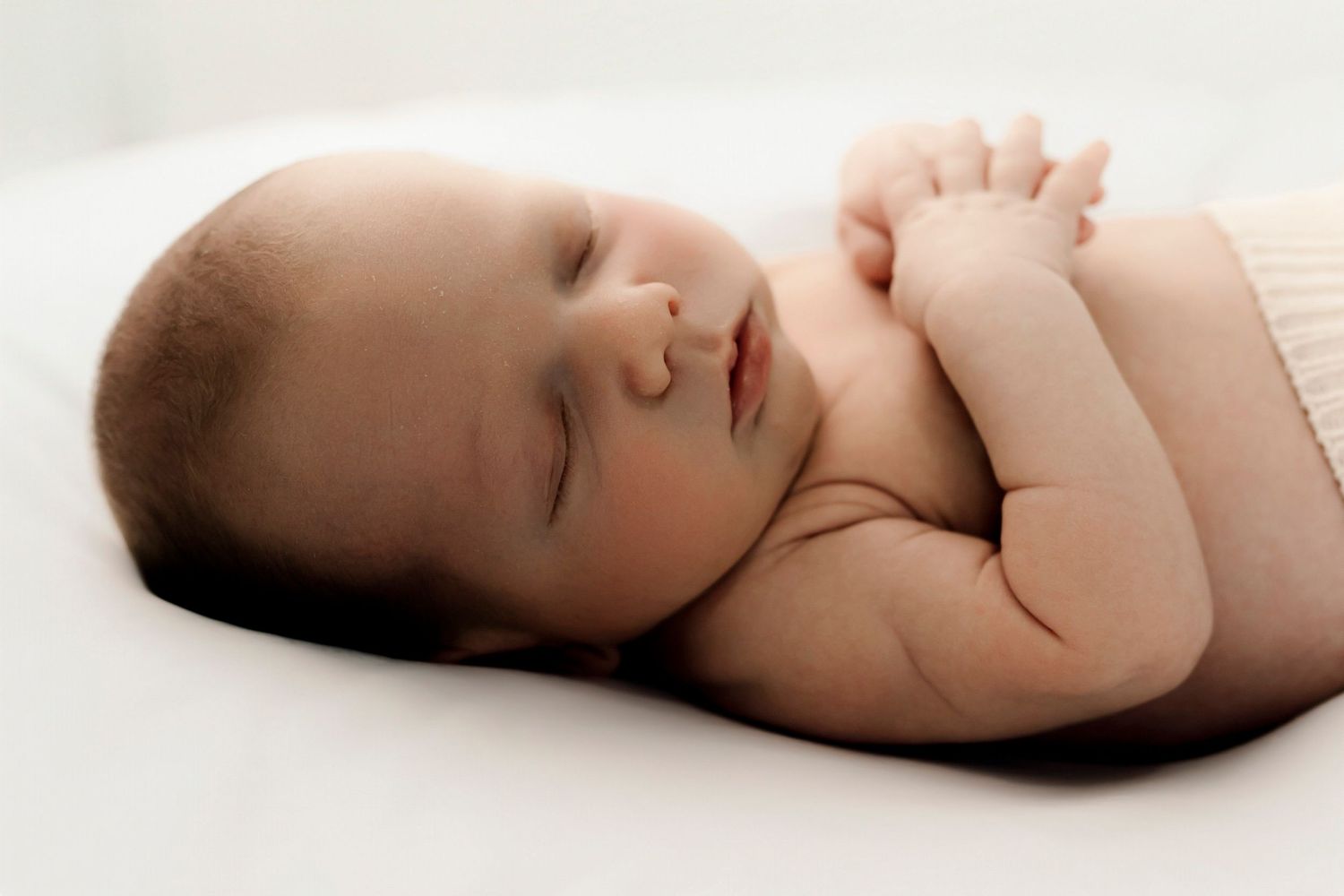Peaceful newborn baby sleeping on white background with tiny hands tucked under chin in serene portrait.
