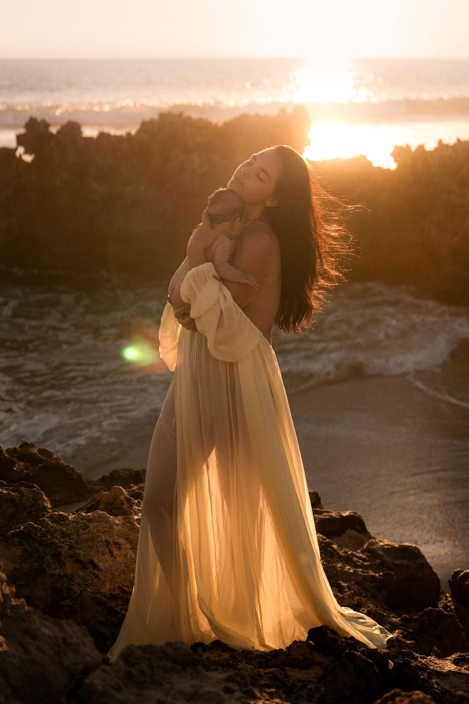 Woman in flowing gown holds baby on rocky shore at golden sunset.