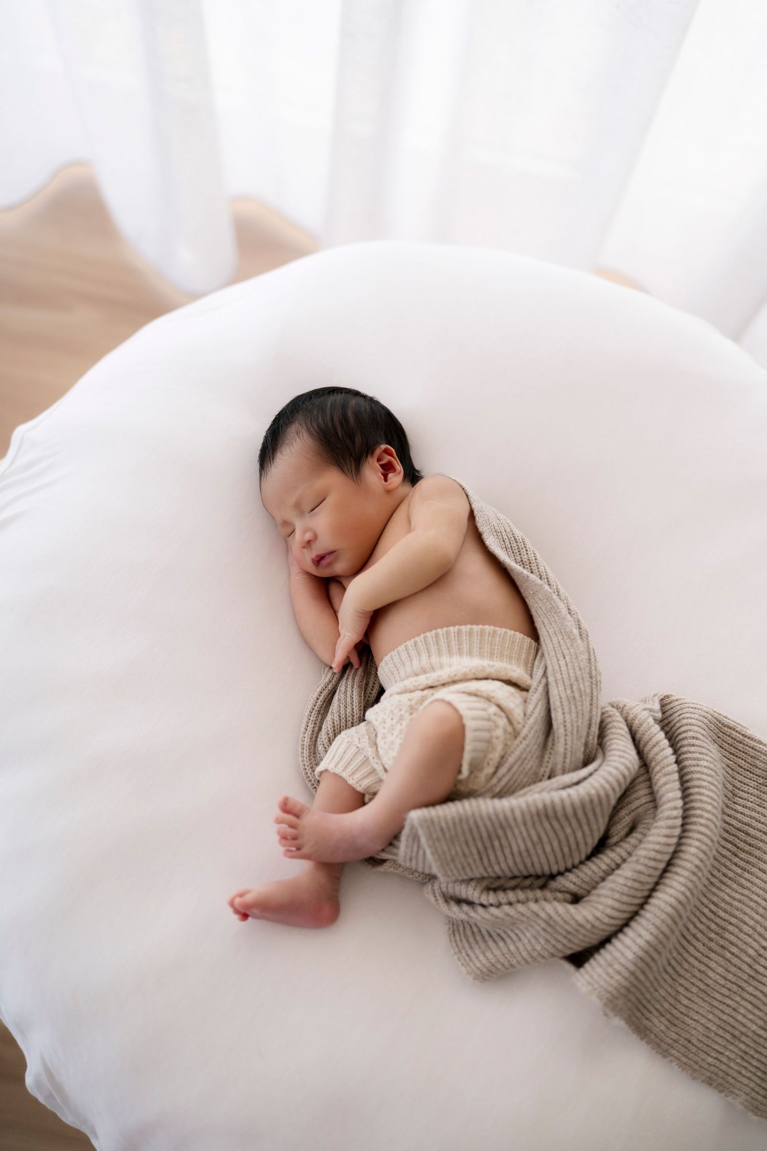 A sleeping newborn baby wrapped in a soft beige knit blanket lies on white fabric.