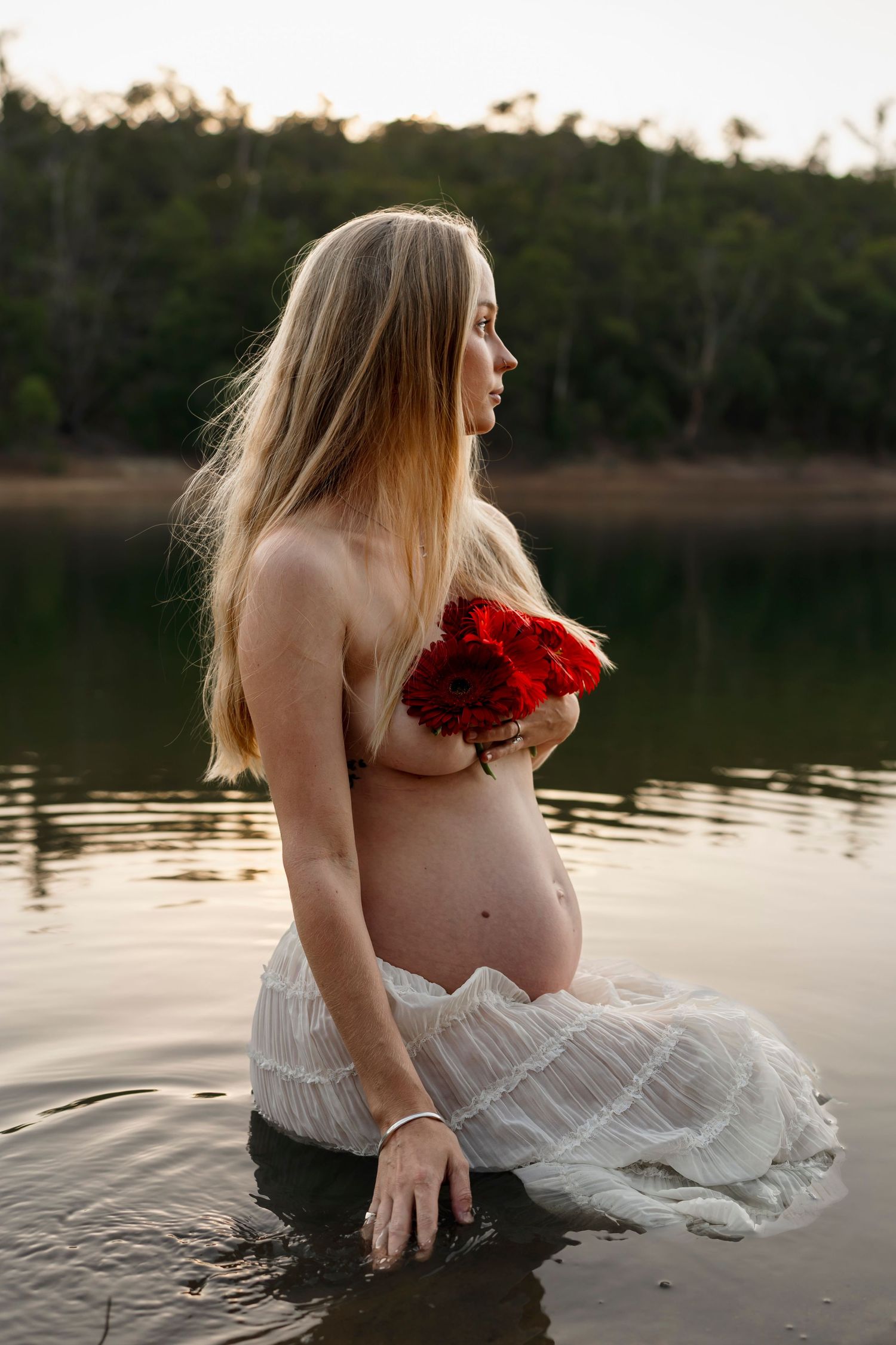Pregnant woman in white dress holding red roses while standing in lake water during golden hour.